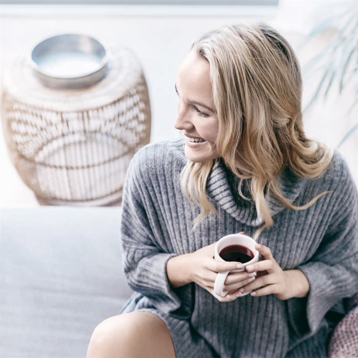 Eine Frau mit blonden Haaren lächelt, während sie eine Tasse Kaffee hält, auf einem Sofa sitzt.