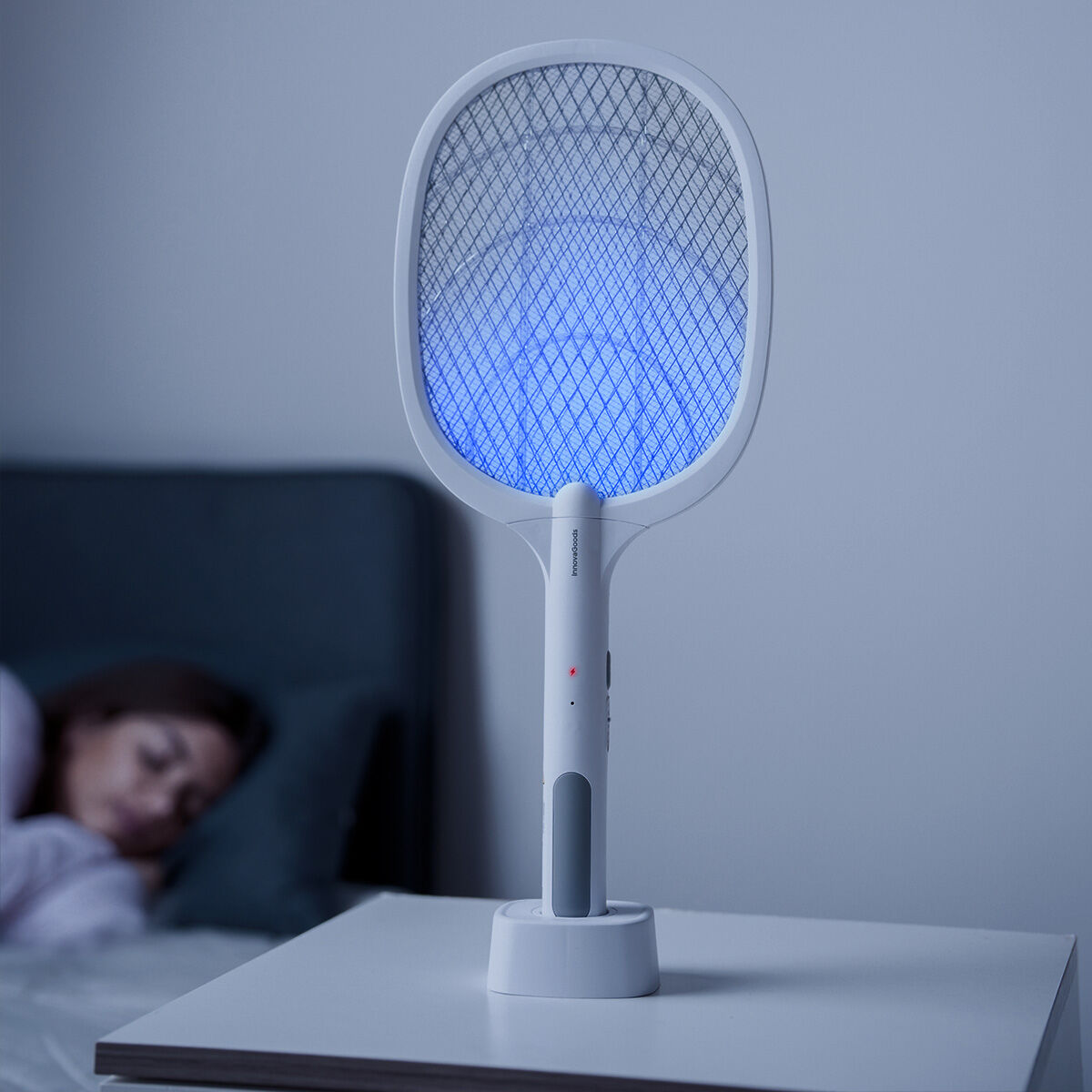 Una racchetta elettrica bianca per zanzare, con bagliore blu, su un comodino accanto a una persona che dorme.
