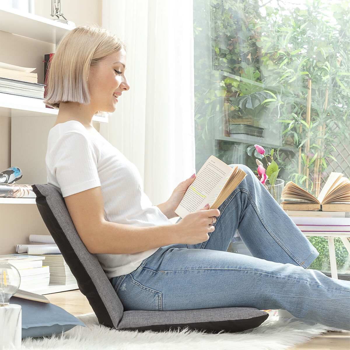 Una mujer sentada en una silla, leyendo un libro. Hay una ventana y una estantería en el fondo.