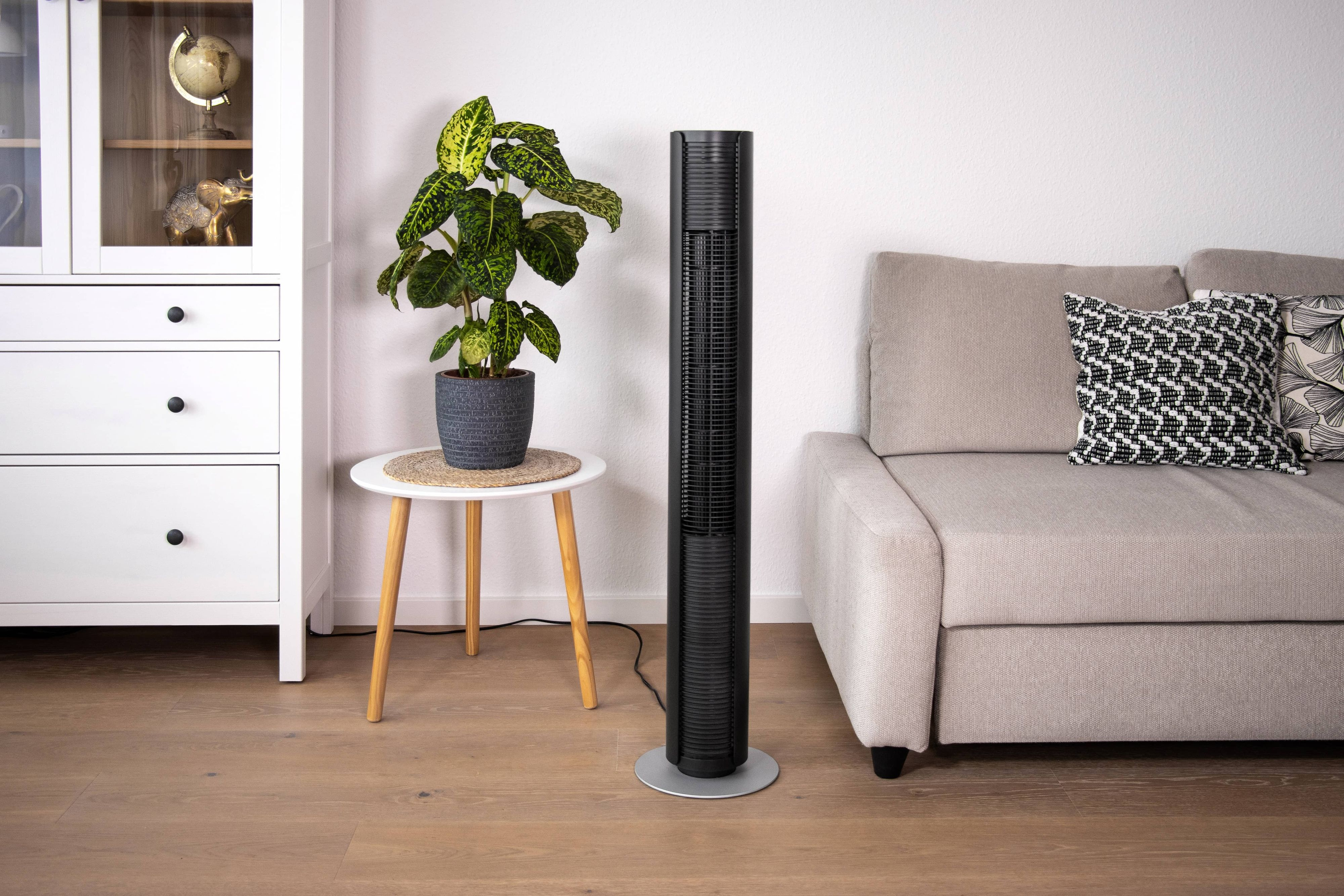 Un ventilateur tour noir près d'une plante, une table et un canapé beige dans une pièce lumineuse.