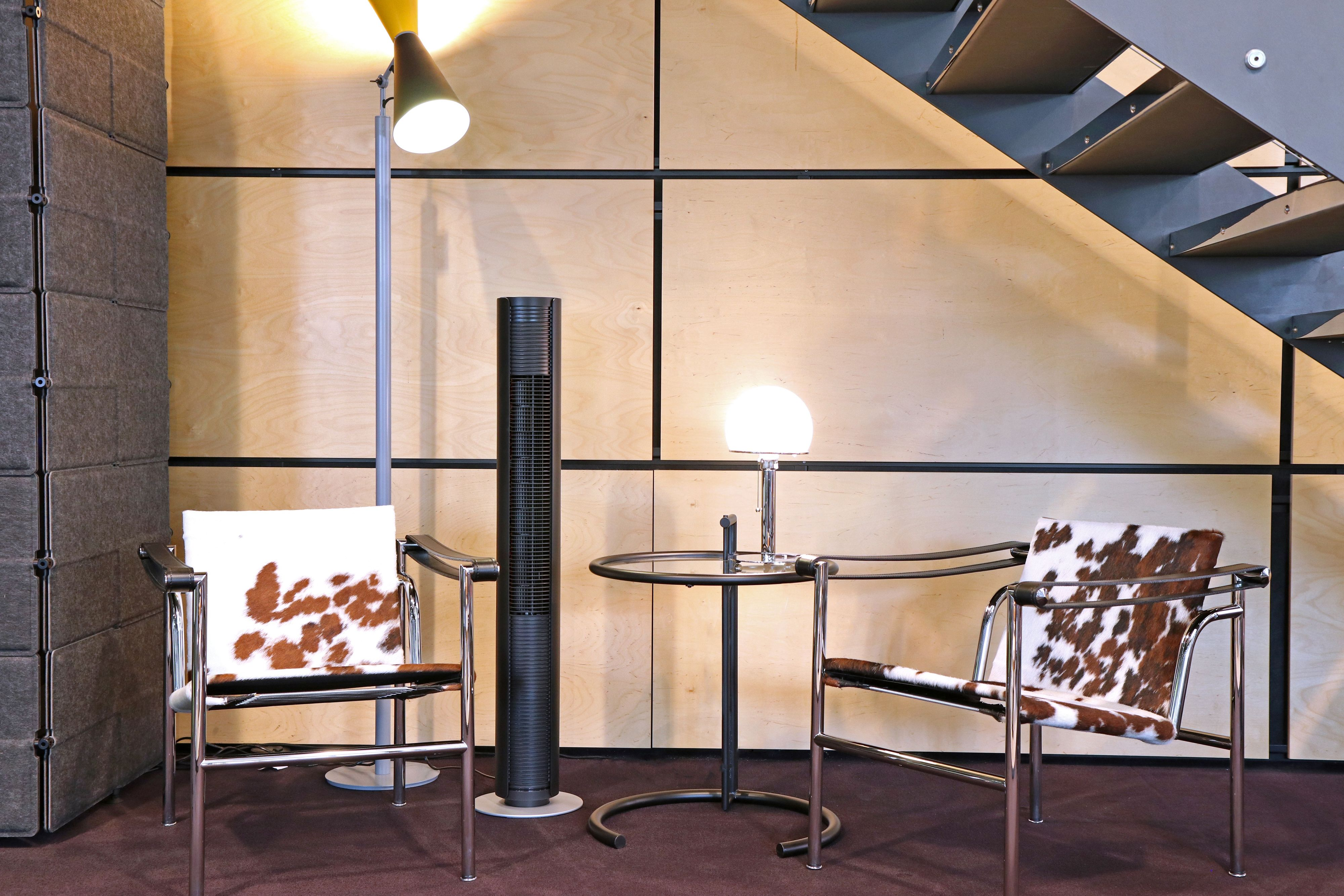 Un ventilateur tour noir entre deux chaises et une petite table, près d'un escalier et d'un mur en bois.