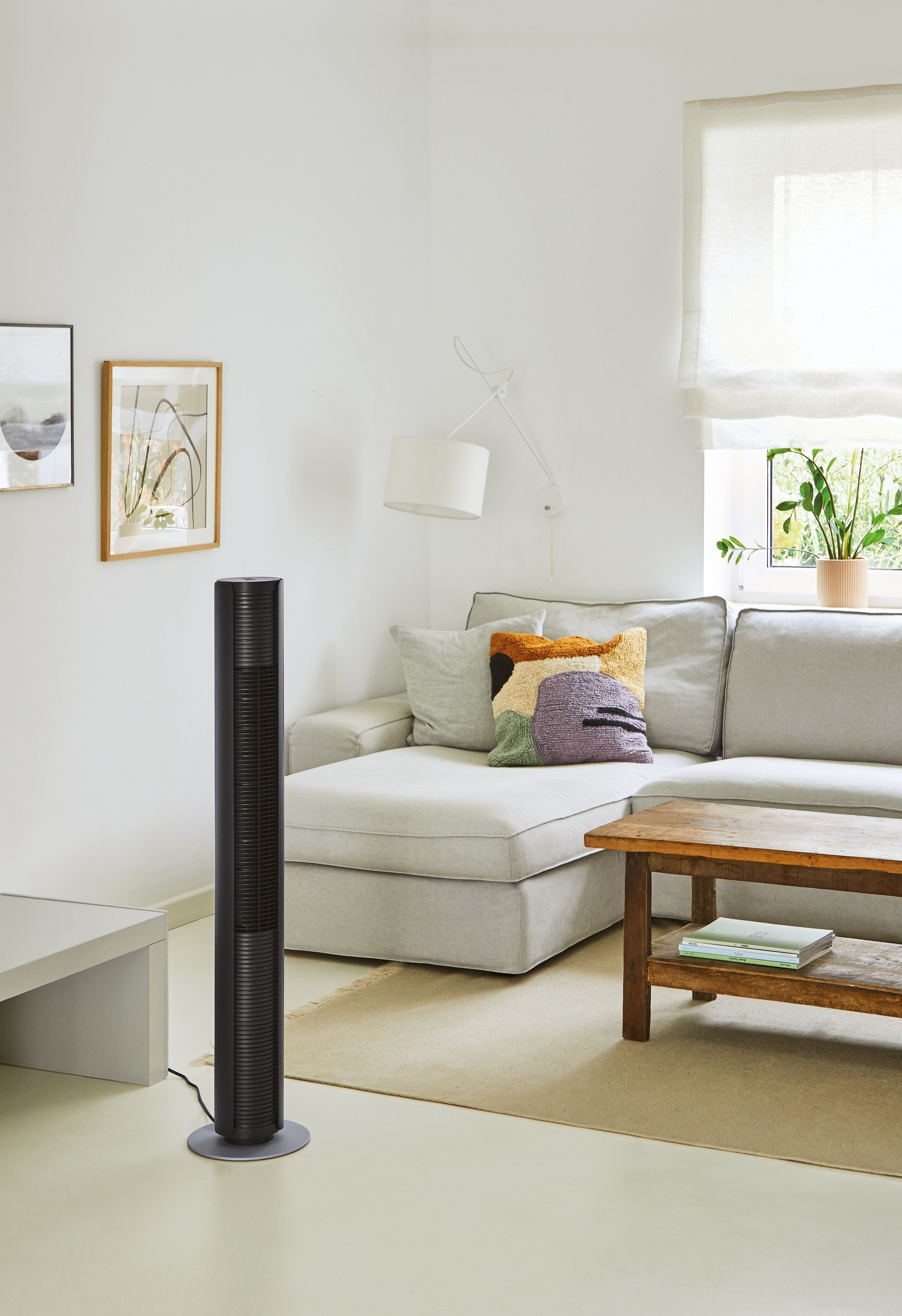 Un ventilateur tour noir dans un salon avec un canapé, une table d'appoint et des œuvres d'art au mur.