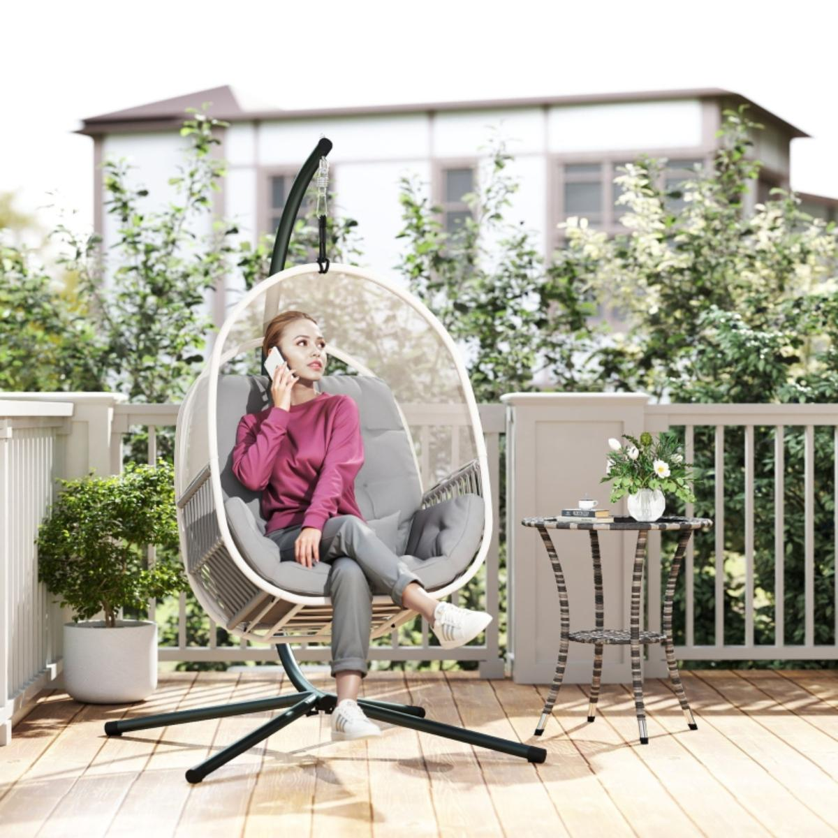 Eine Frau in einem rosa Hemd sitzt in einem hängenden Eierstuhl auf einem Deck und telefoniert. Ein Tisch steht neben ihr.