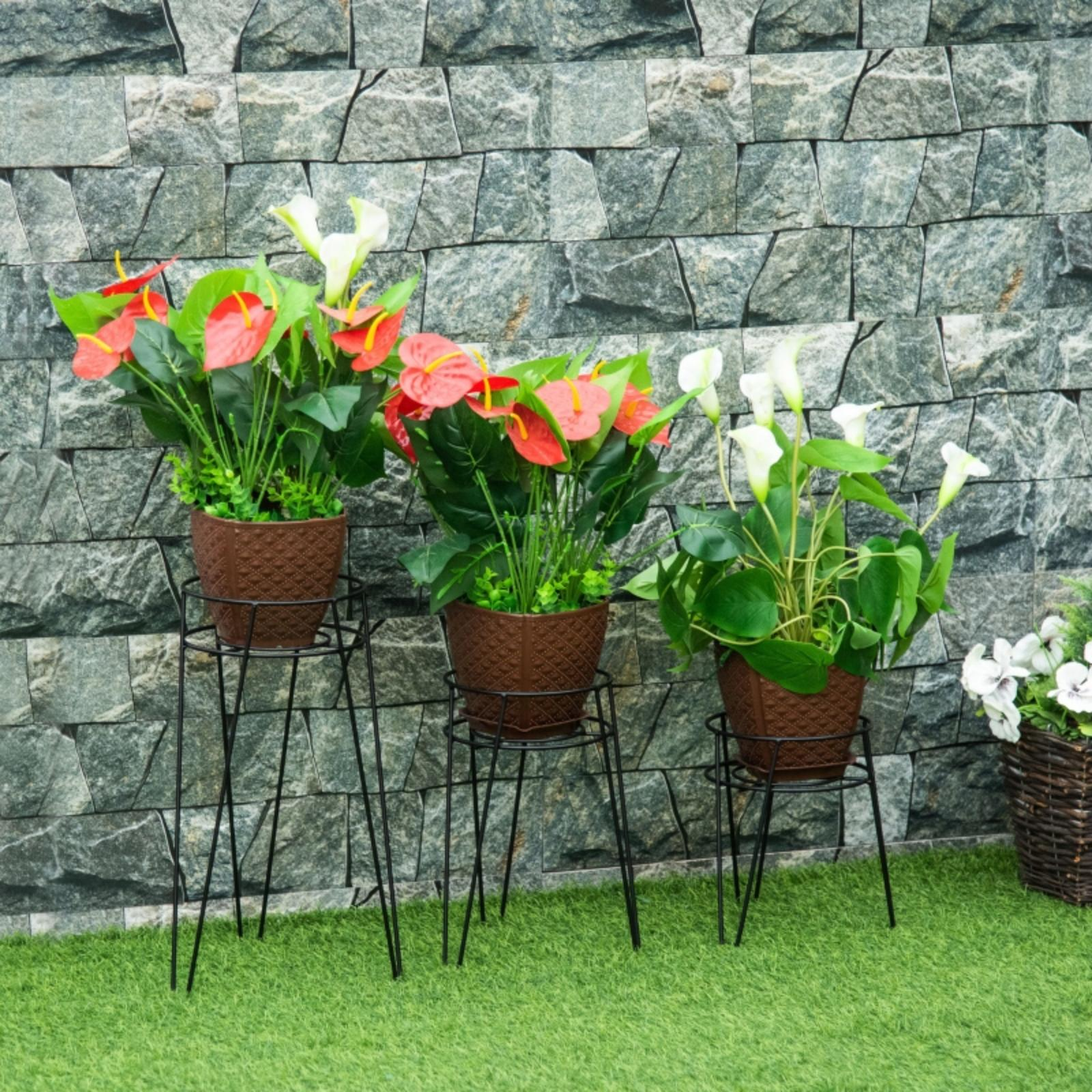 Drei Topfpflanzen mit roten und weißen Blüten stehen auf schwarzen Ständern vor einer Steinmauer.