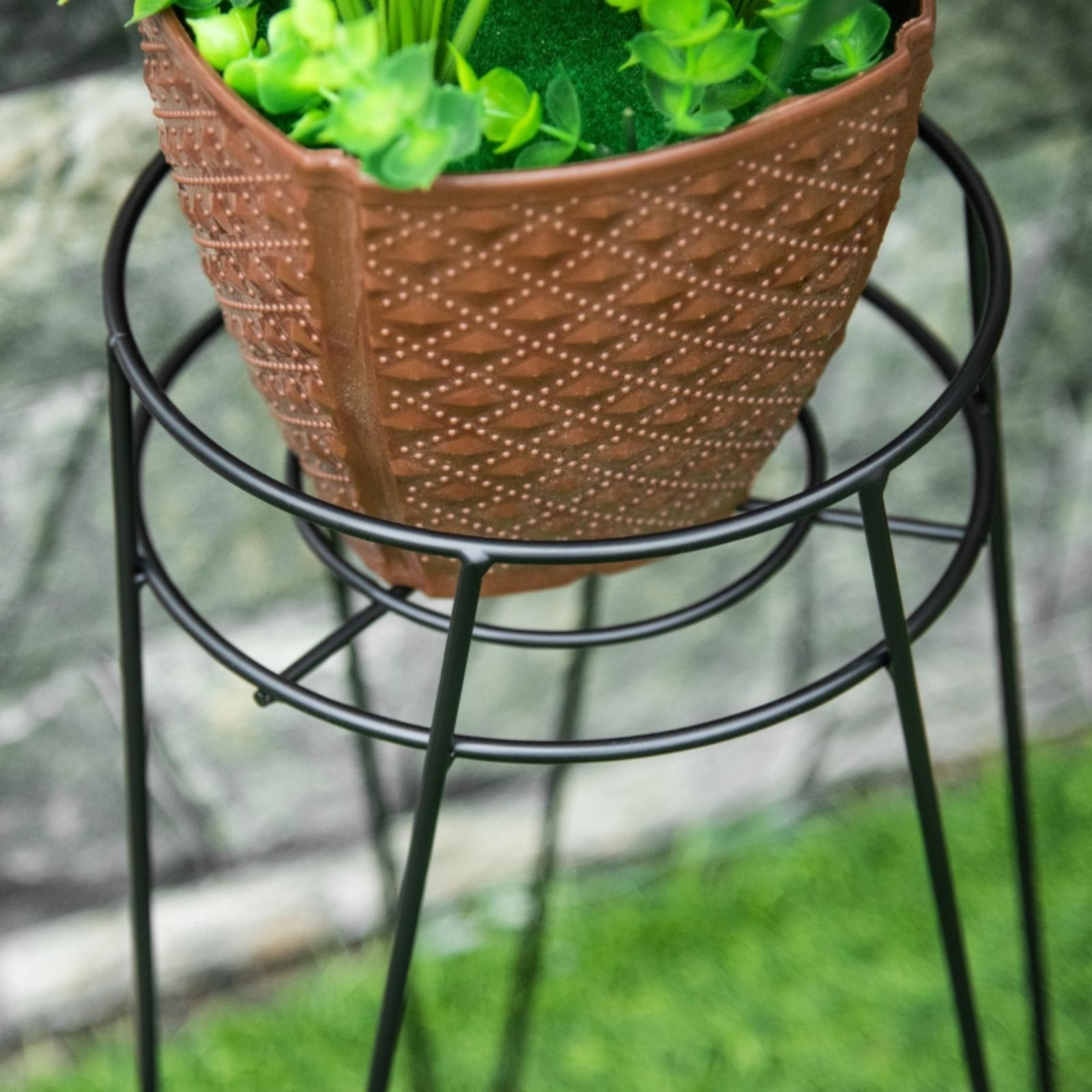 Ein Blumentopf auf einem schwarzen Metallständer. Der Topf ist braun mit einem gemusterten Design. Der Hintergrund ist verschwommenes grünes Gras.