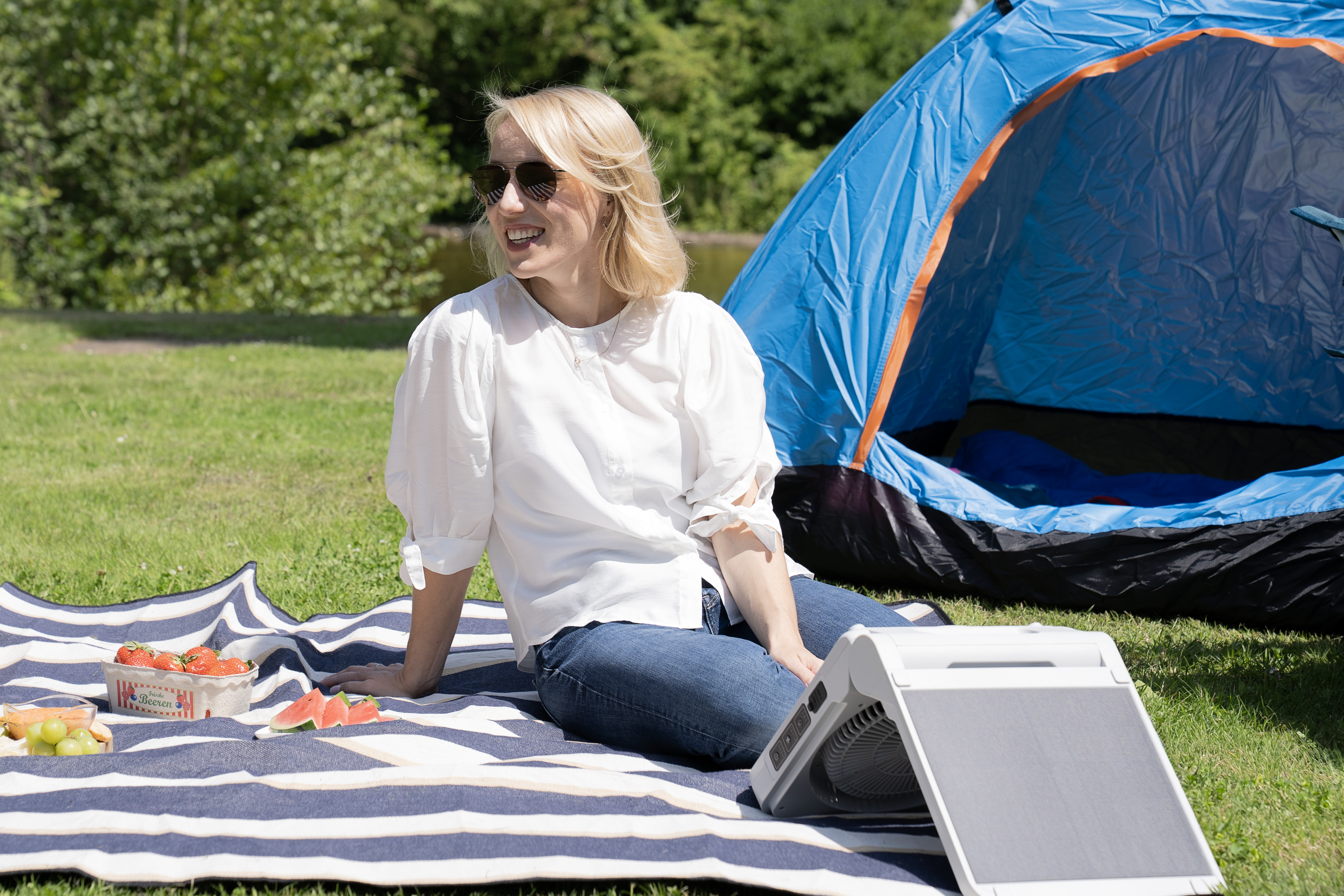 Kobieta siedzi na kocu w pobliżu namiotu, uśmiecha się, z przedmiotami piknikowymi i wentylatorem w pobliżu.