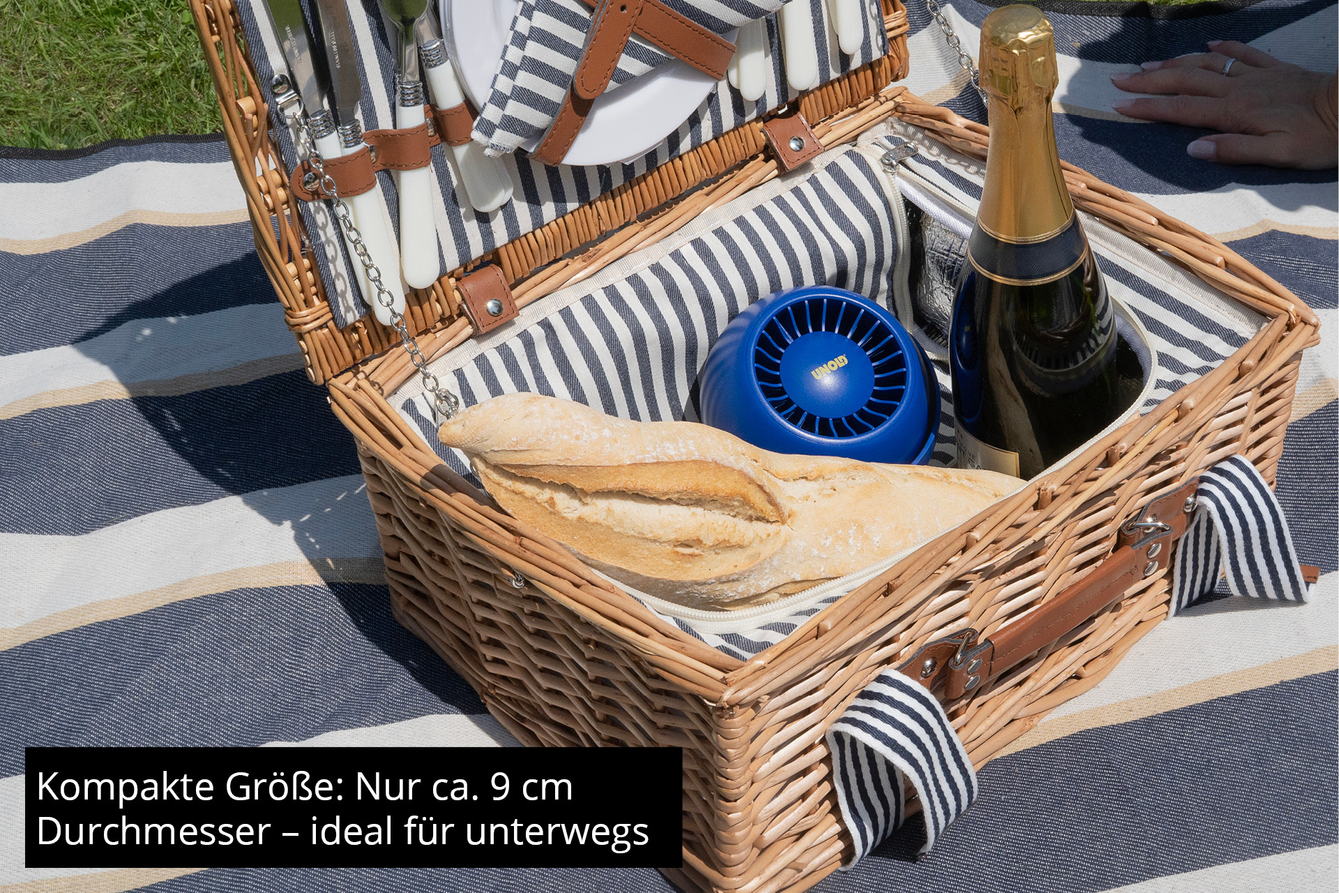 Cesto da picnic in vimini con cibo e un piccolo ventilatore blu all'interno. Bottiglia di champagne visibile.