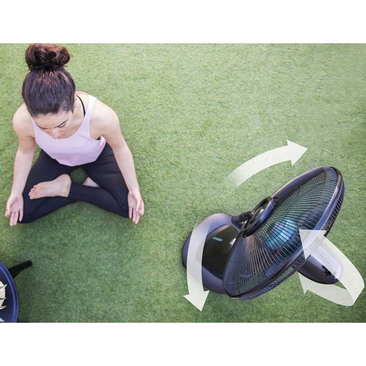 Una mujer sentada en hierba verde cerca de un ventilador negro. Flechas indican el movimiento giratorio.