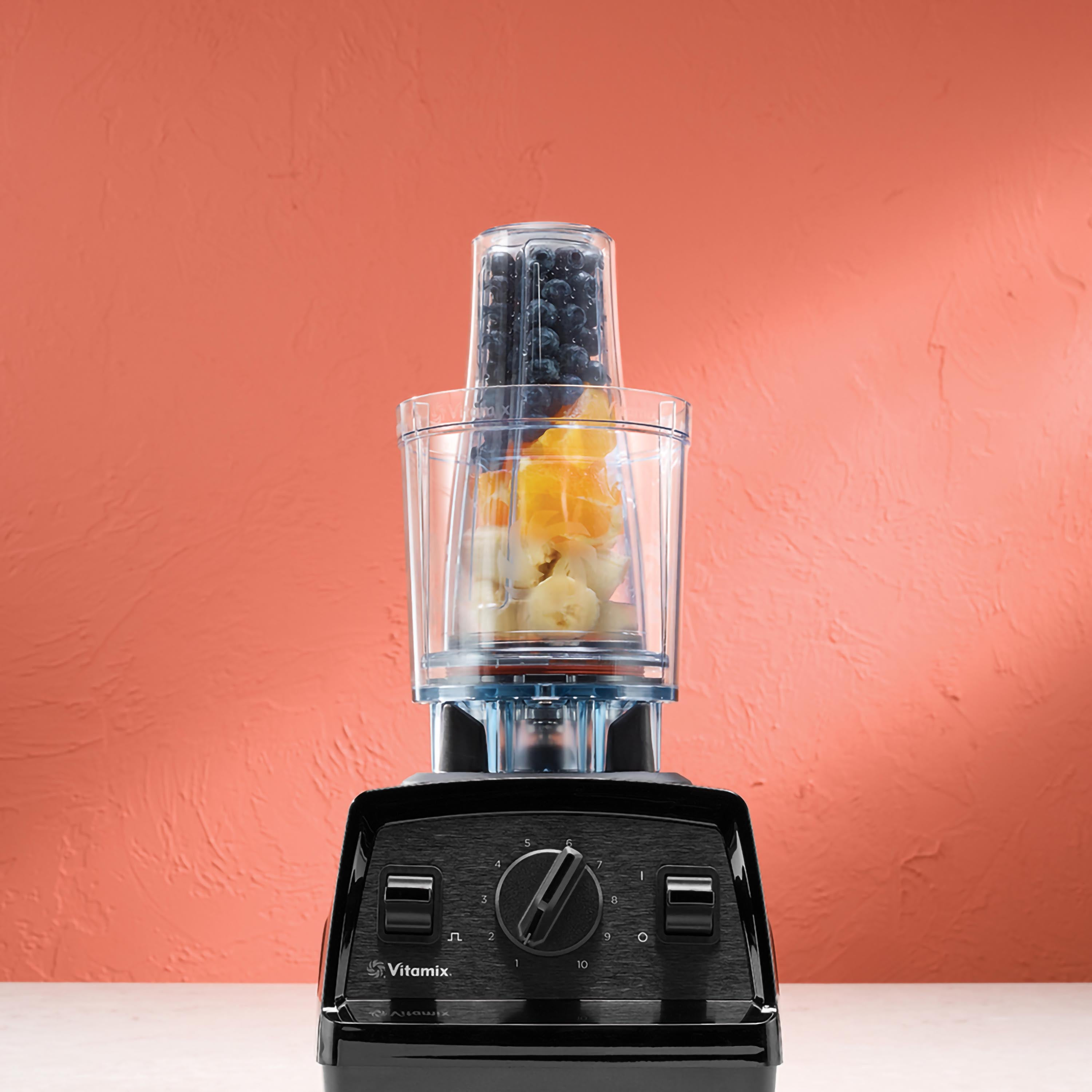 Ein schwarzer Vitamix-Mixer mit einem mit Früchten gefüllten Behälter. Im Hintergrund eine rote Wand.