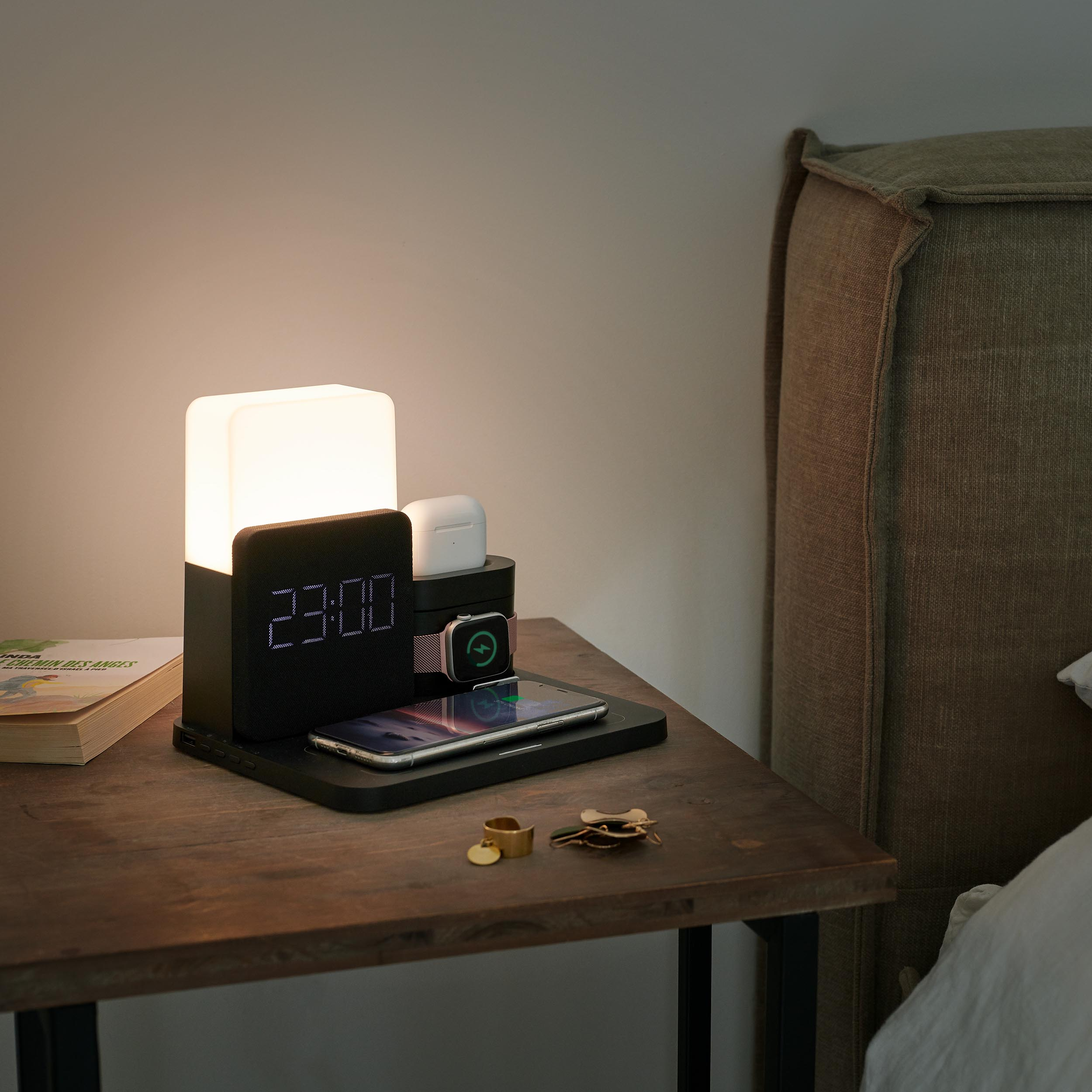 Czarna stacja ładująca z zegarem cyfrowym, telefonem, zegarkiem, słuchawkami i lampą. Drewniany stół.