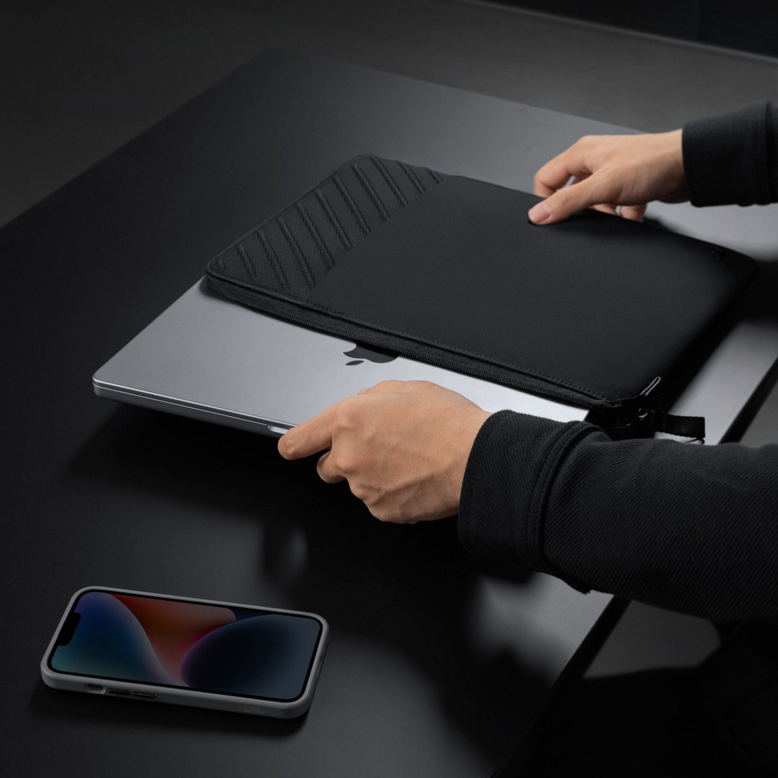 Dłonie otwierające czarną torbę na laptopa na czarnym stole, z telefonem na pierwszym planie.