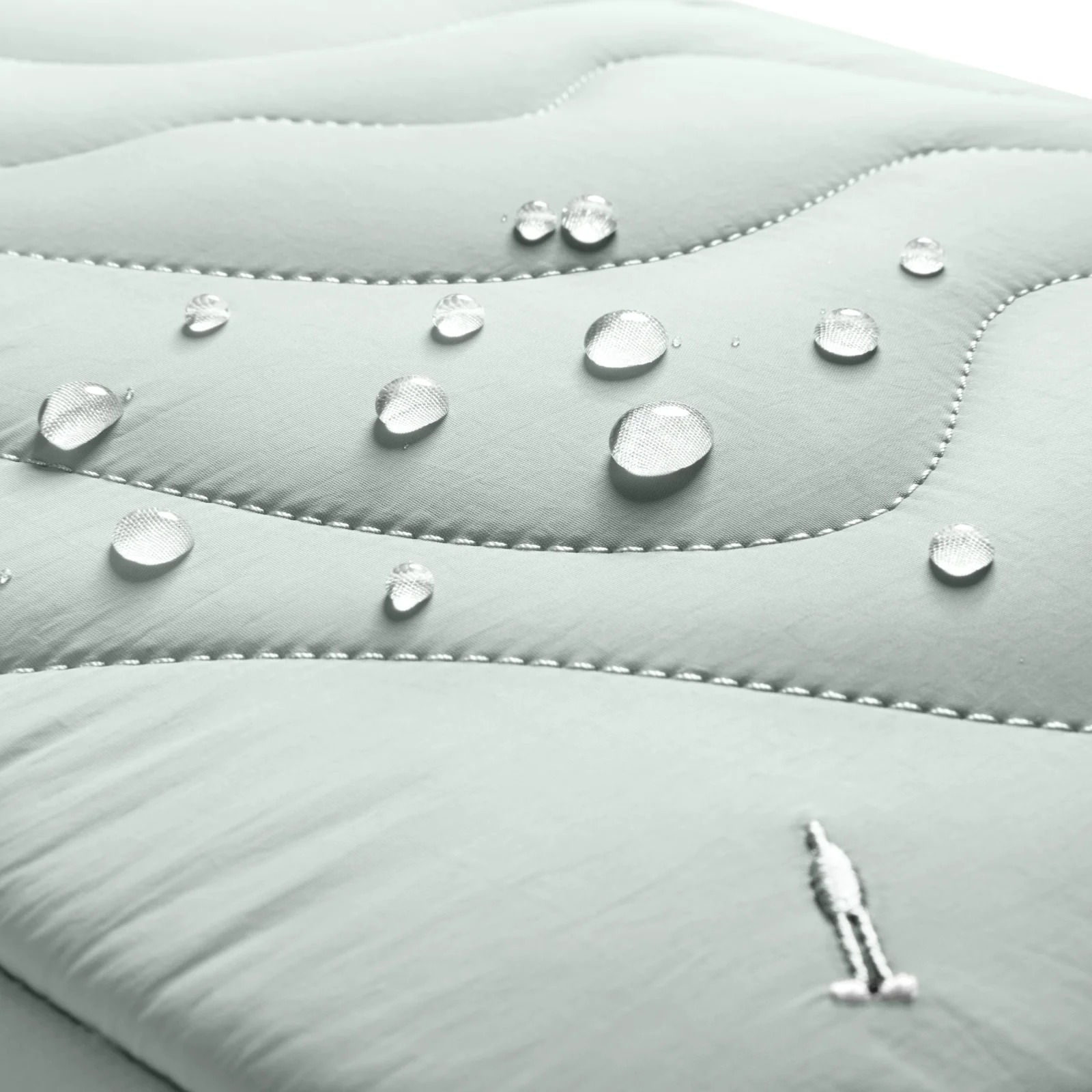 Zbliżenie jasnozielonej pikowanej tkaniny z kroplami wody. Obecne jest małe haftowane logo.