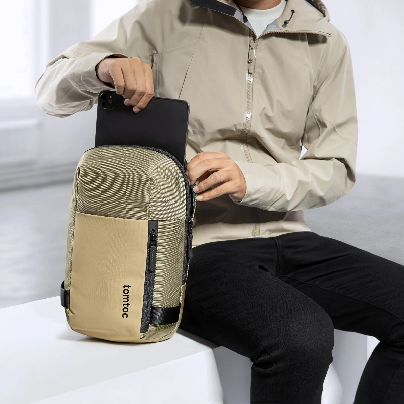 Osoba wkładająca tablet do beżowej torby na ramię tomtoc. Osoba ma na sobie beżową kurtkę.