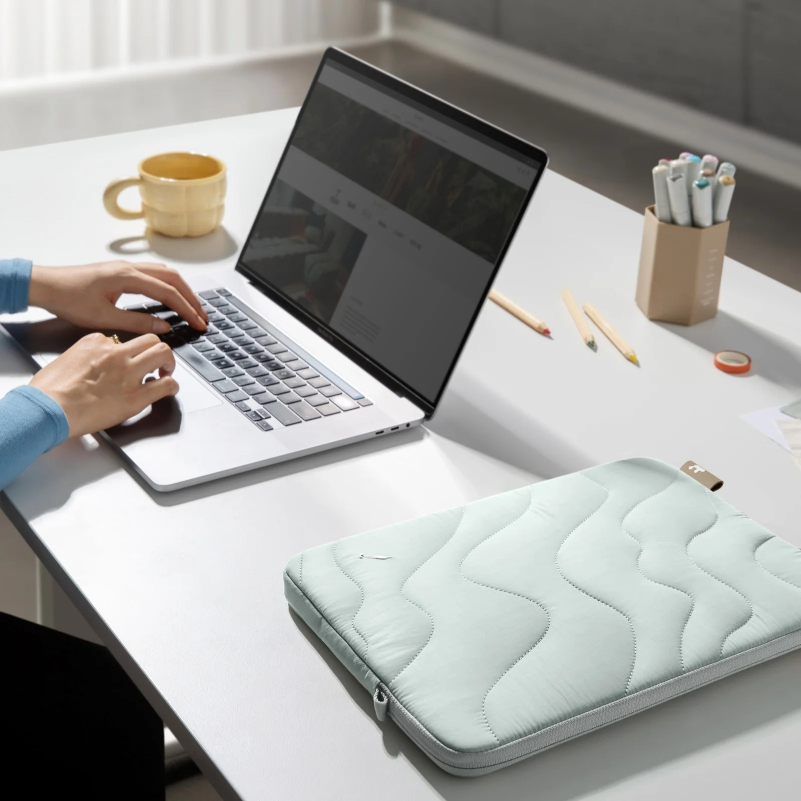 Osoba pisze na laptopie na białym biurku. Obok znajduje się jasnozielone etui na laptopa.