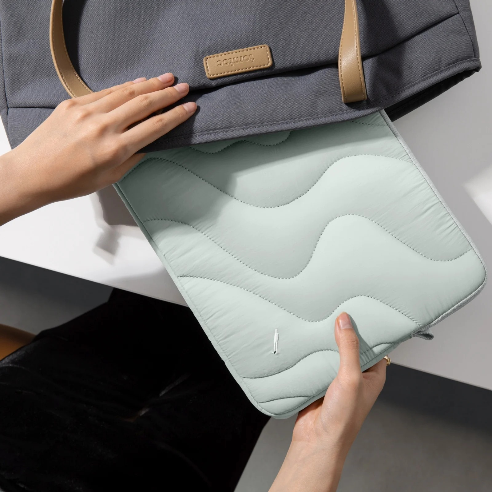 Osoba wkłada jasnozielony pokrowiec na laptopa do szarej torby typu tote. Torba ma brązową skórzaną metkę.