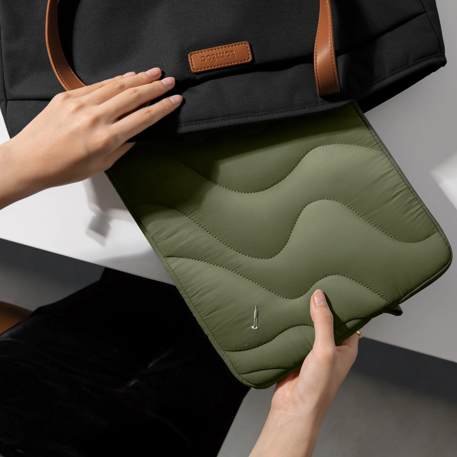 Ręce trzymające zieloną pikowaną torbę na laptopa. Czarna torba w tle na białej powierzchni.