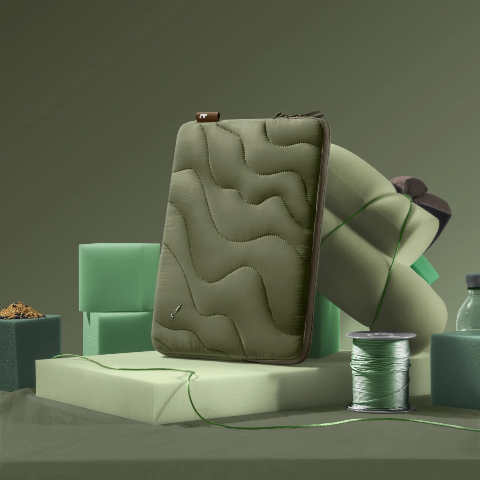 Zielona torba na laptopa w kolorze oliwkowym. Leży na zielonej powierzchni z innymi przedmiotami. Tło jest zielone.