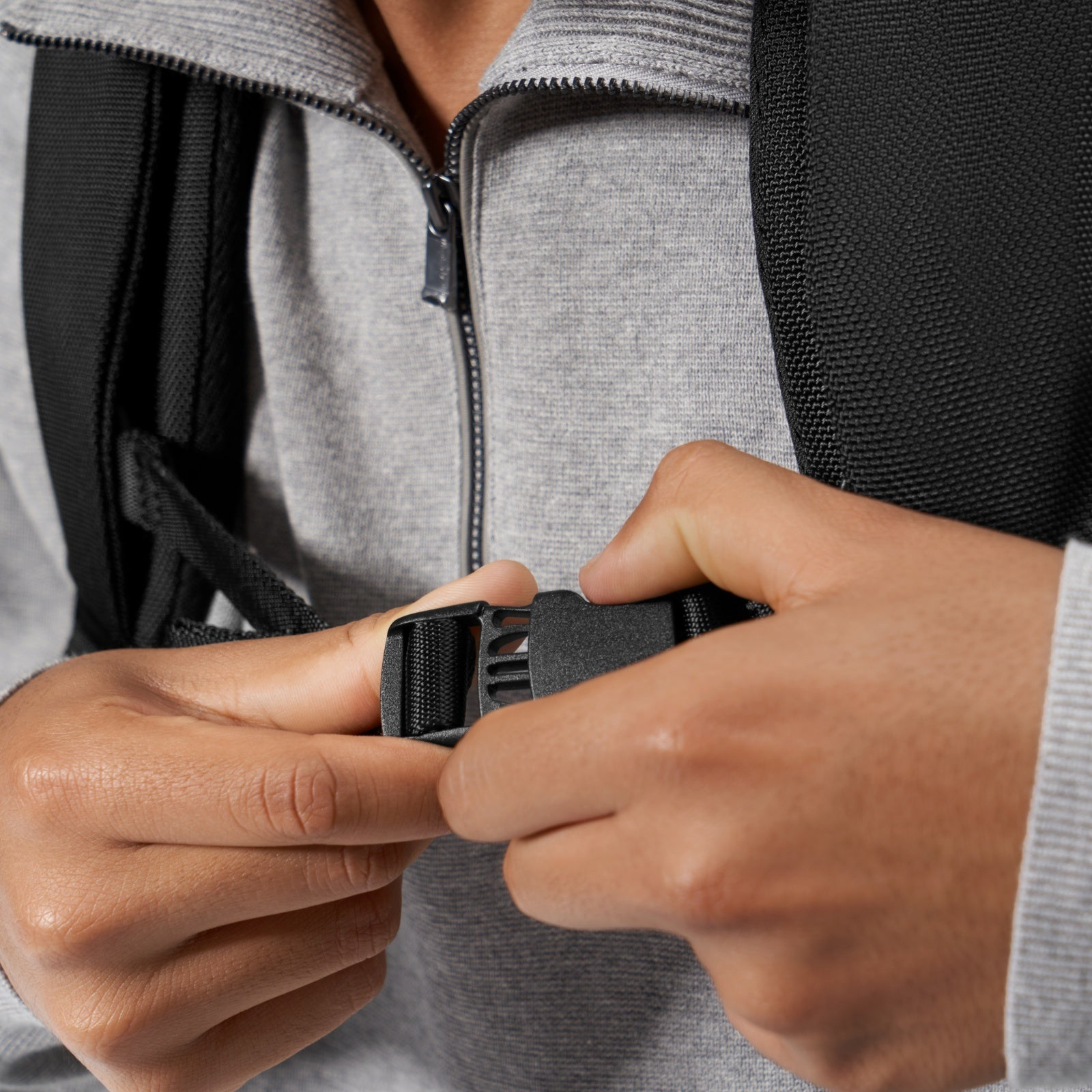 Zbliżenie dłoni zapinających czarną klamrę na pasku plecaka, w szarym swetrze.