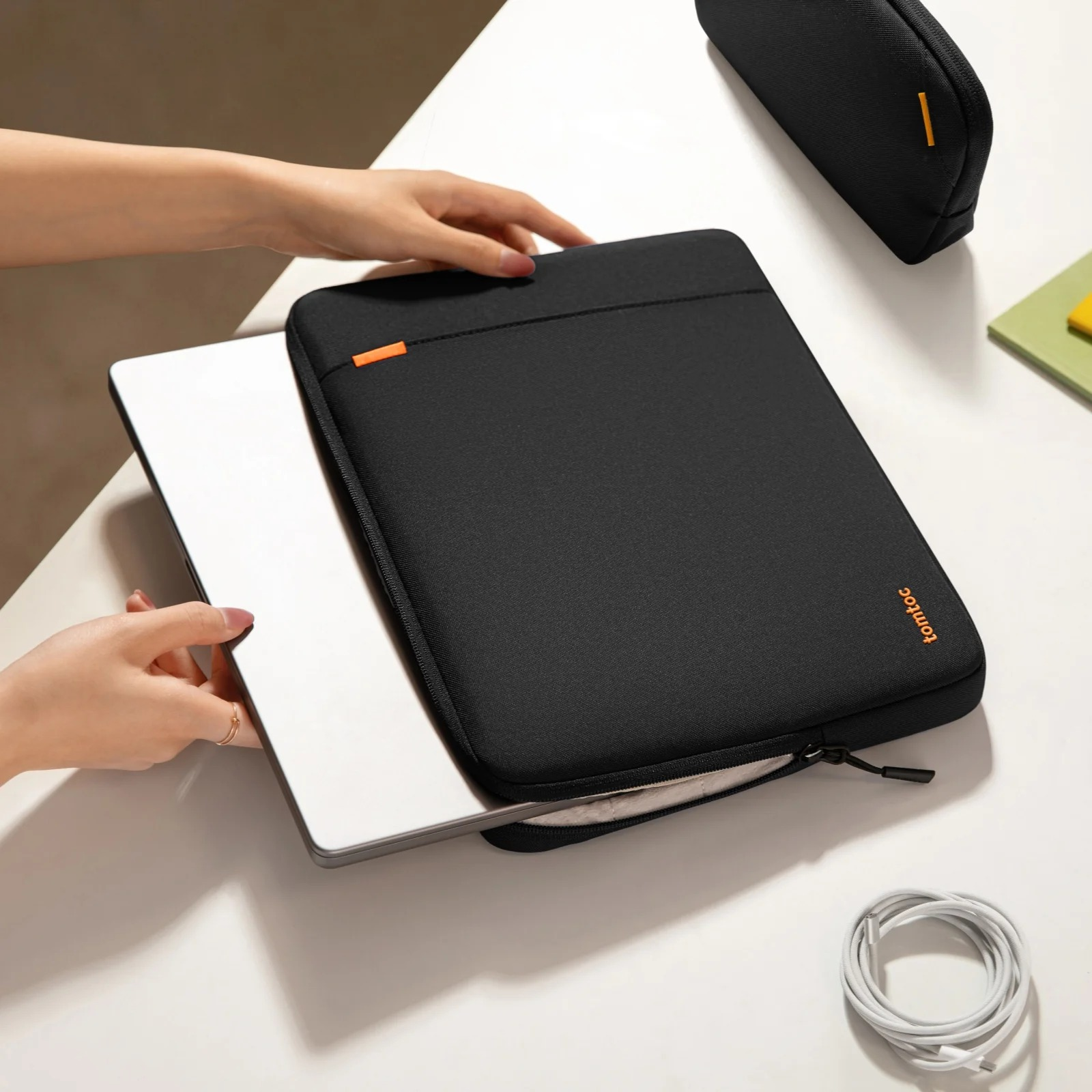 Ręce wkładają laptopa do czarnej obudowy na białej powierzchni. Widoczna jest kolejna obudowa i kabel.