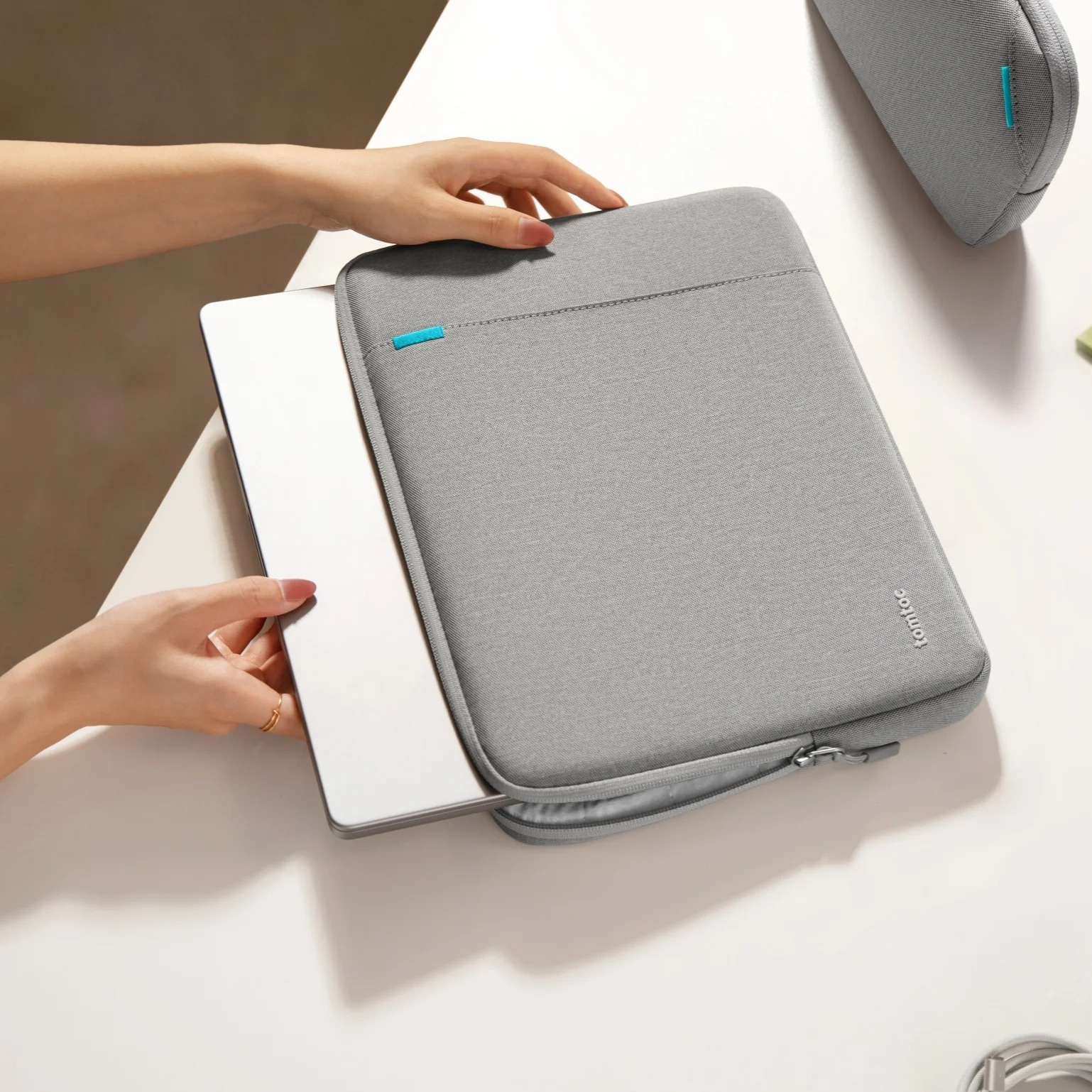 Ręce wkładają laptopa do szarej obudowy na białej powierzchni. Widoczny jest również kabel.