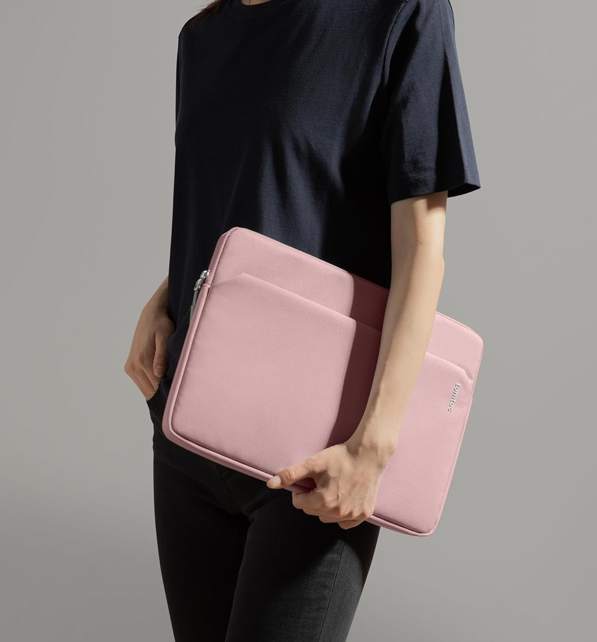 Kobieta trzymająca różową torbę na laptopa. Czarny t-shirt, spodnie. Szare tło.