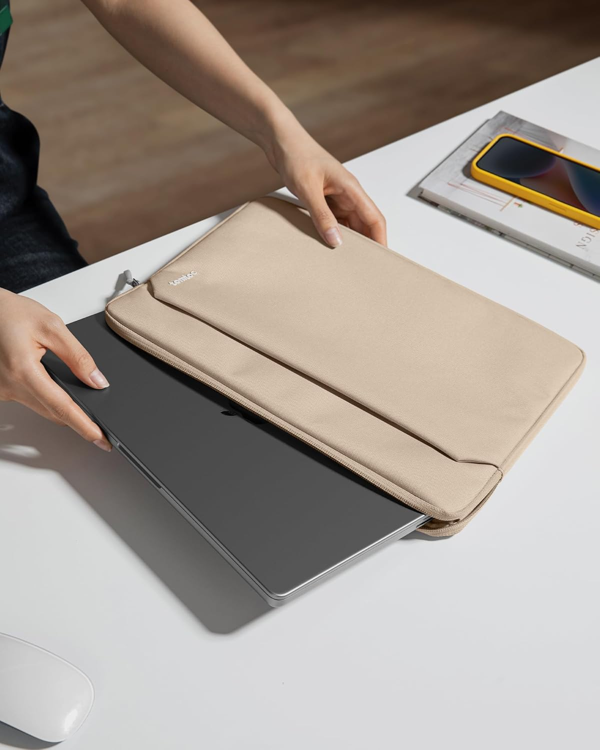 Ręce wkładające laptopa do beżowej torby. Biały stół, książki i telefon również widoczne.