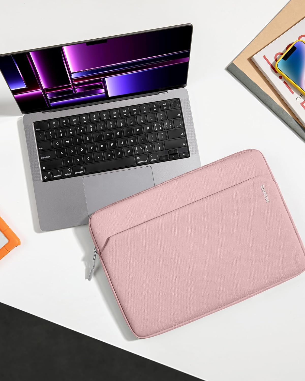 Laptop i różowa torba na białym stole. Książki i telefon również na stole.