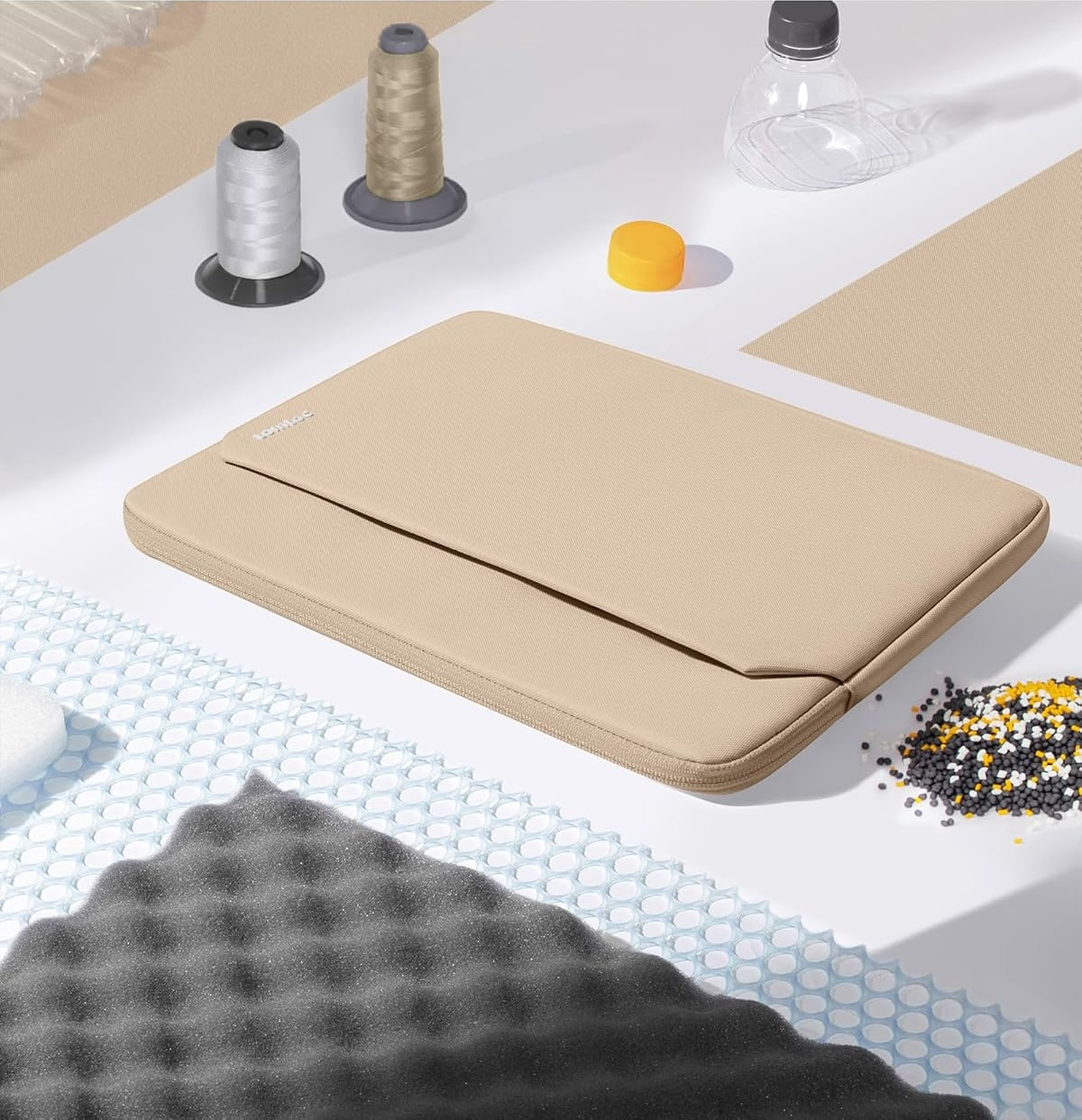 Beżowa torba na laptopa z akcesoriami na białej powierzchni. Zawiera nici, butelkę wody i piankę.