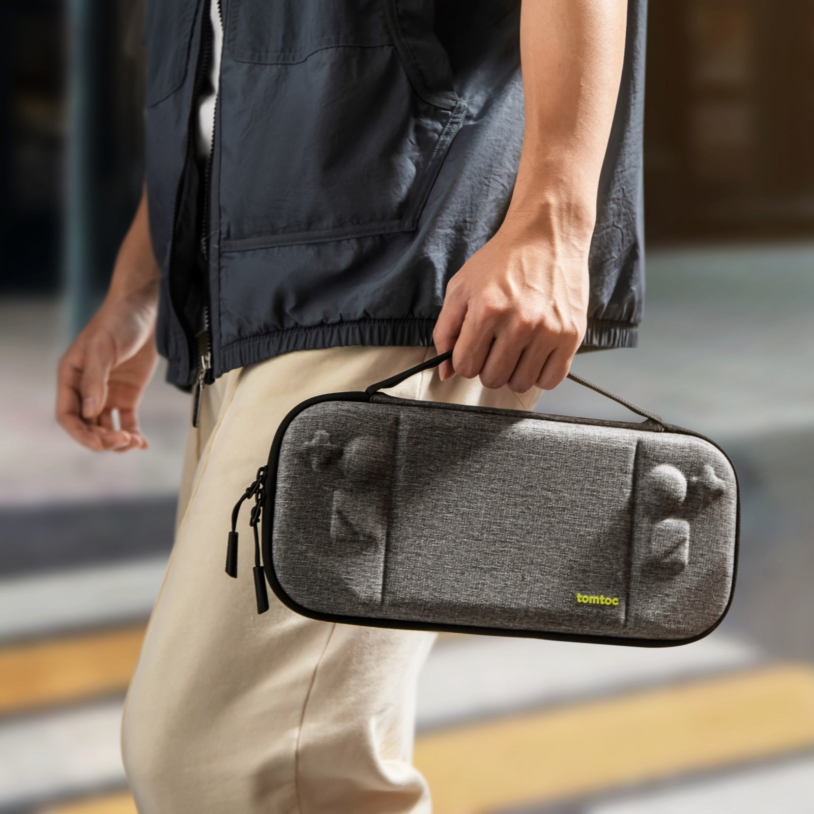 Osoba trzyma szarą walizkę z uchwytem. Walizka ma kształt konsoli do gier.