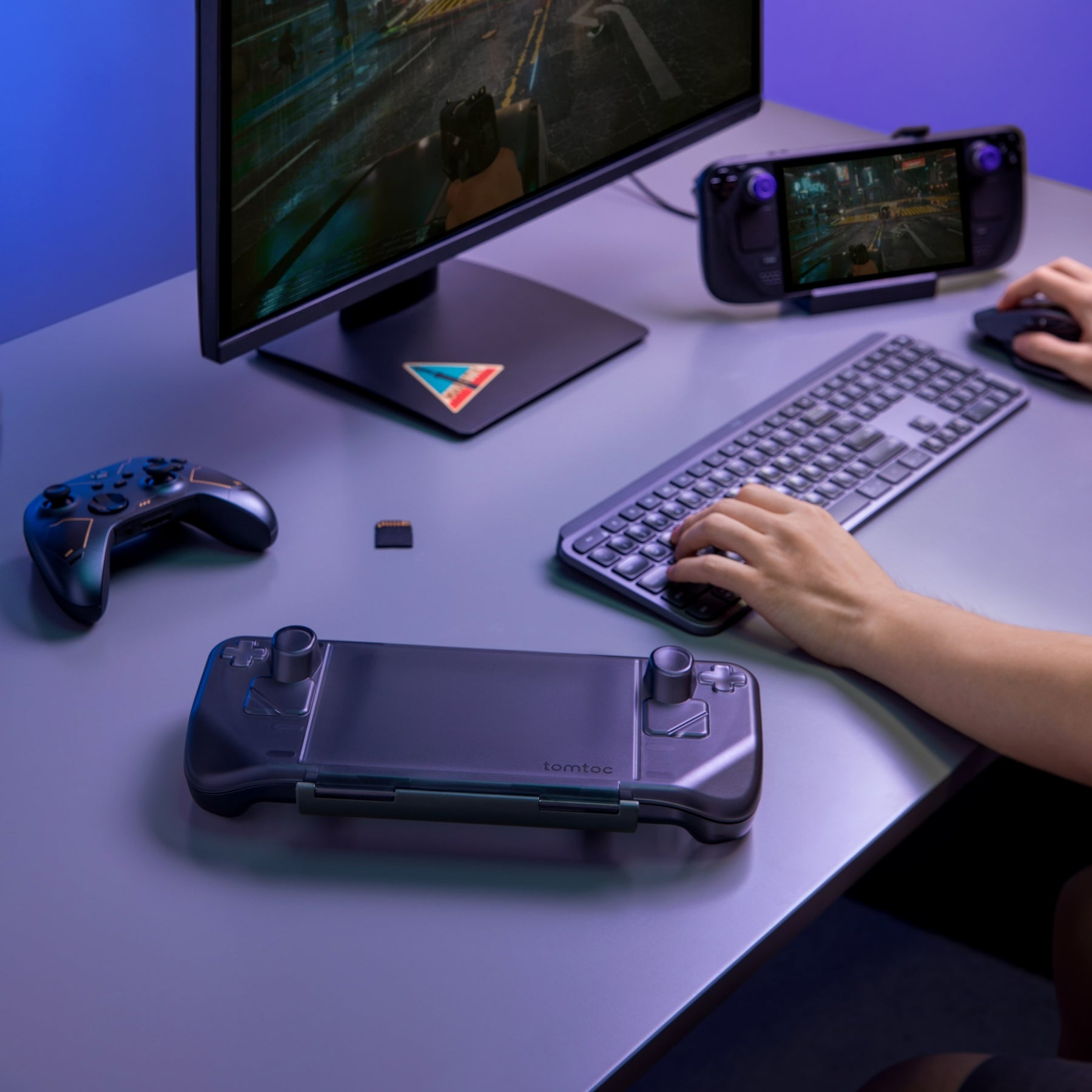 Konfiguracja do gier z dwiema konsolami przenośnymi, monitorem, klawiaturą i kontrolerem na szarym biurku.