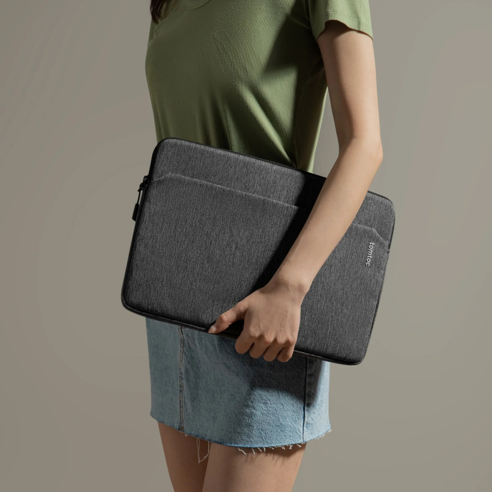 Kobieta trzyma szarą torbę na laptopa. Torba ma kieszeń i zamek błyskawiczny. Ma na sobie zieloną koszulę.