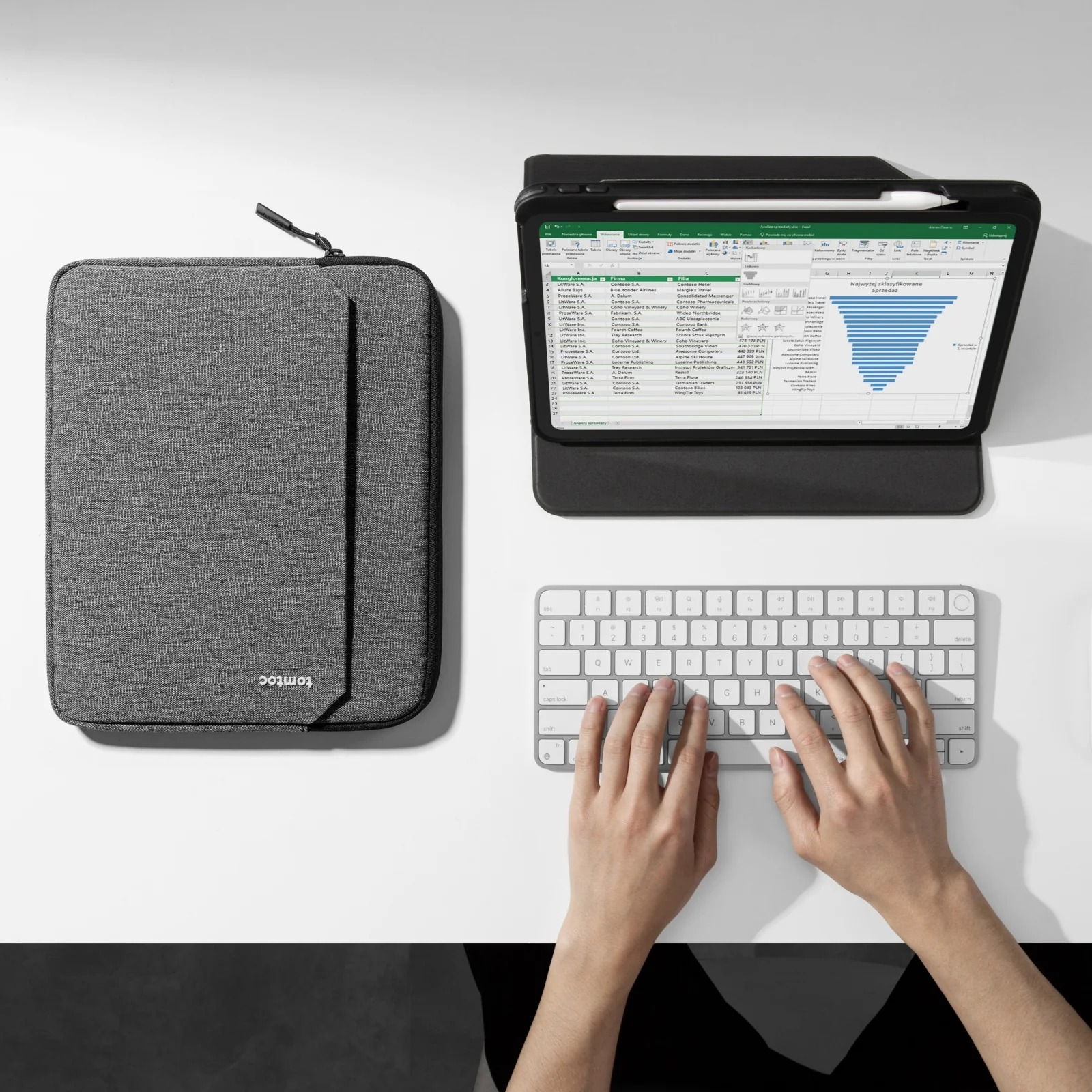 Laptop, tablet i klawiatura znajdują się na białym stole. Szara torba na laptopa leży obok nich. Białe tło.