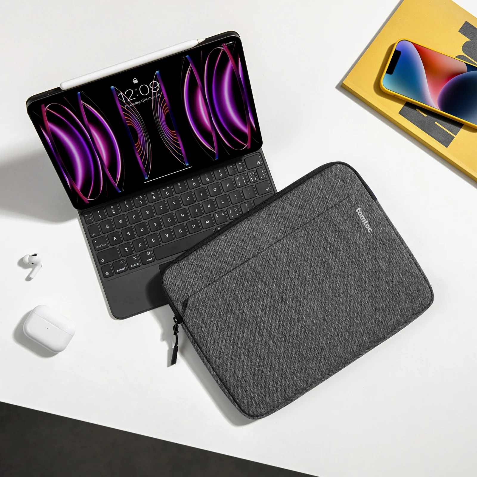 Tablet z klawiaturą na stole. Szare etui i AirPods obok. Żółta książka i długopis na stole.