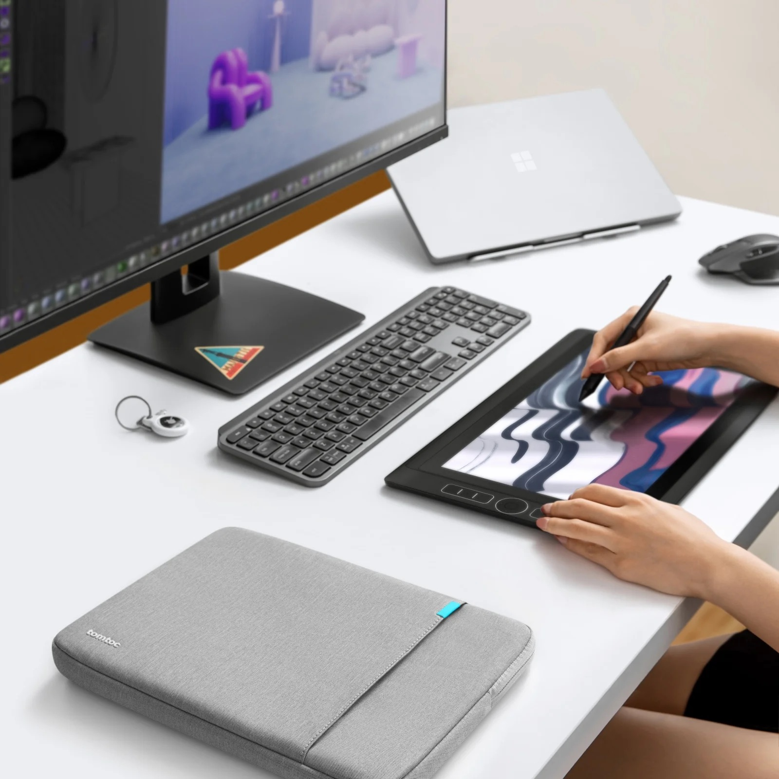 Osoba używa rysika na tablecie. Klawiatura i laptop na białym biurku. Szare etui na biurku.