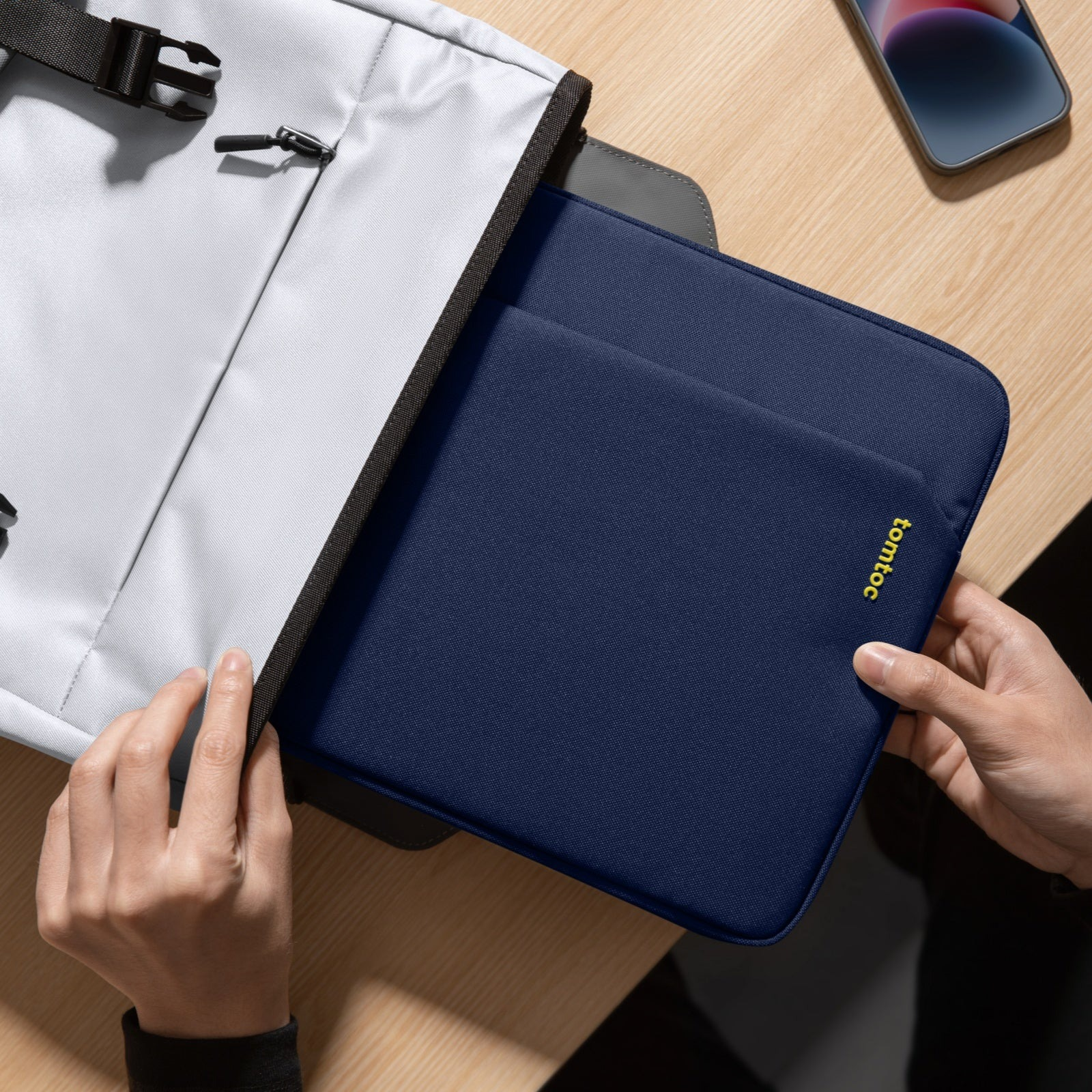 Ręce trzymają granatową torbę na laptopa, częściowo w szarym plecaku. Widoczny telefon.