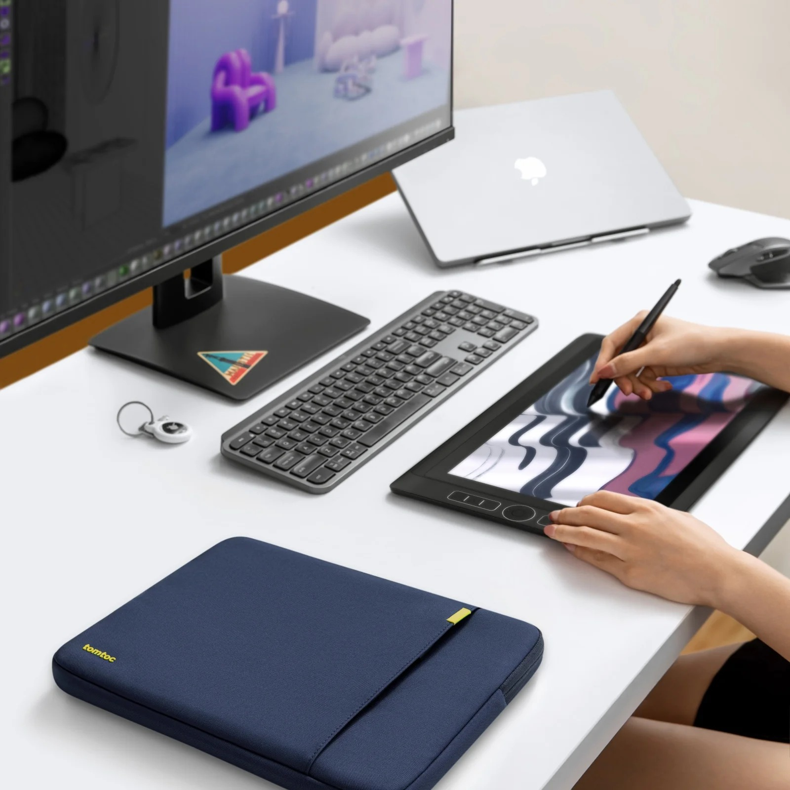 Biurko z monitorem, laptopem, klawiaturą, tabletem piórkowym i granatową torbą na laptopa.