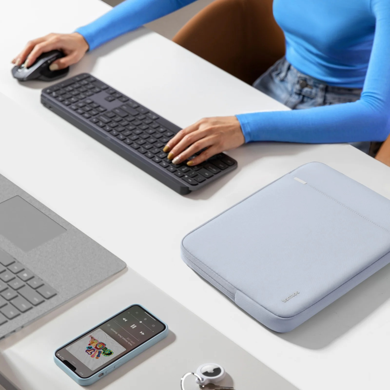 Osoba używająca klawiatury i myszy, z telefonem, etui i laptopem na biurku.