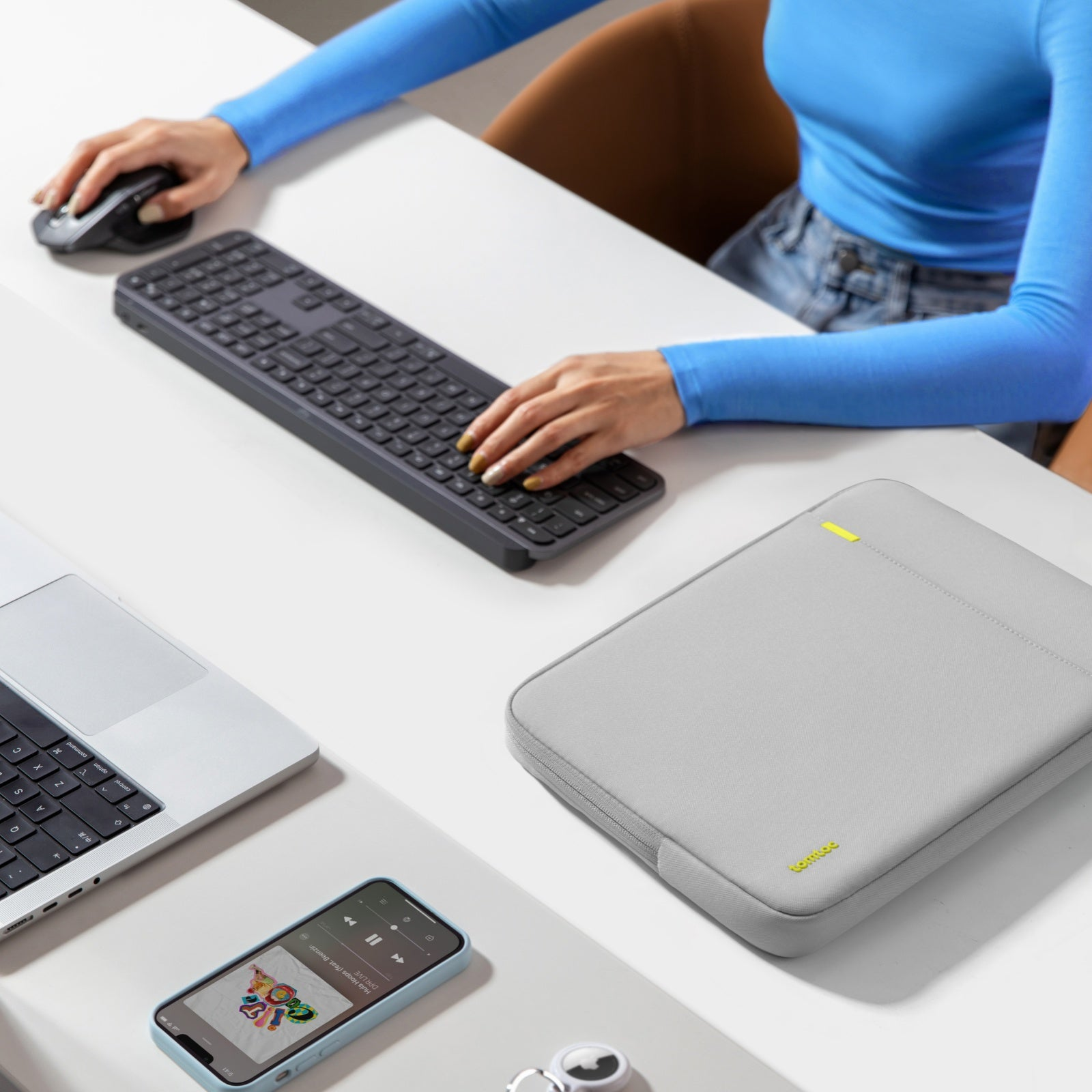 Osoba pracuje przy biurku z laptopem, klawiaturą, telefonem i etui na laptopa.