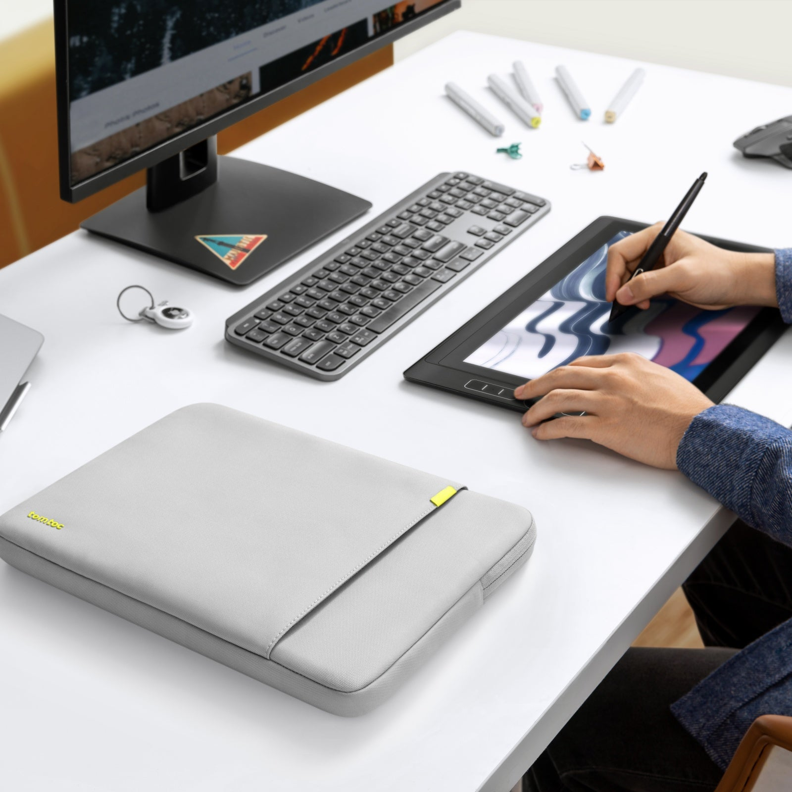 Osoba korzystająca z tabletu i klawiatury na białym biurku. Na biurku znajduje się szara torba na laptopa.