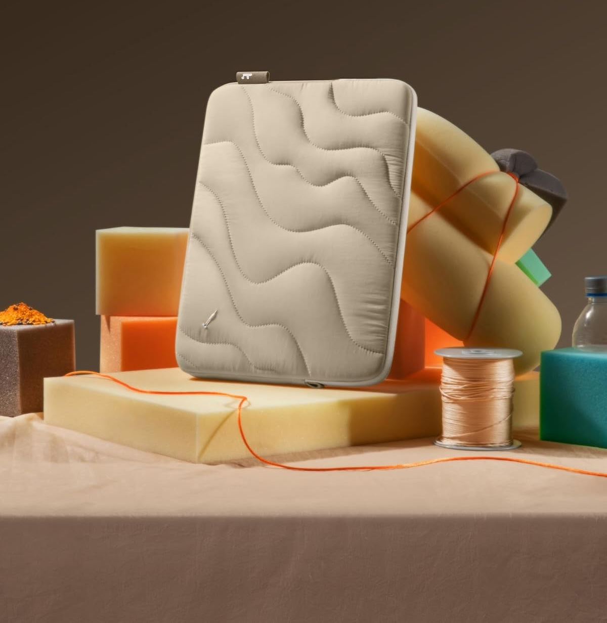 Beżowe pikowane etui na laptopa na blokach piankowych. Pomarańczowa nić i proszek na pierwszym planie. Brązowe tło.