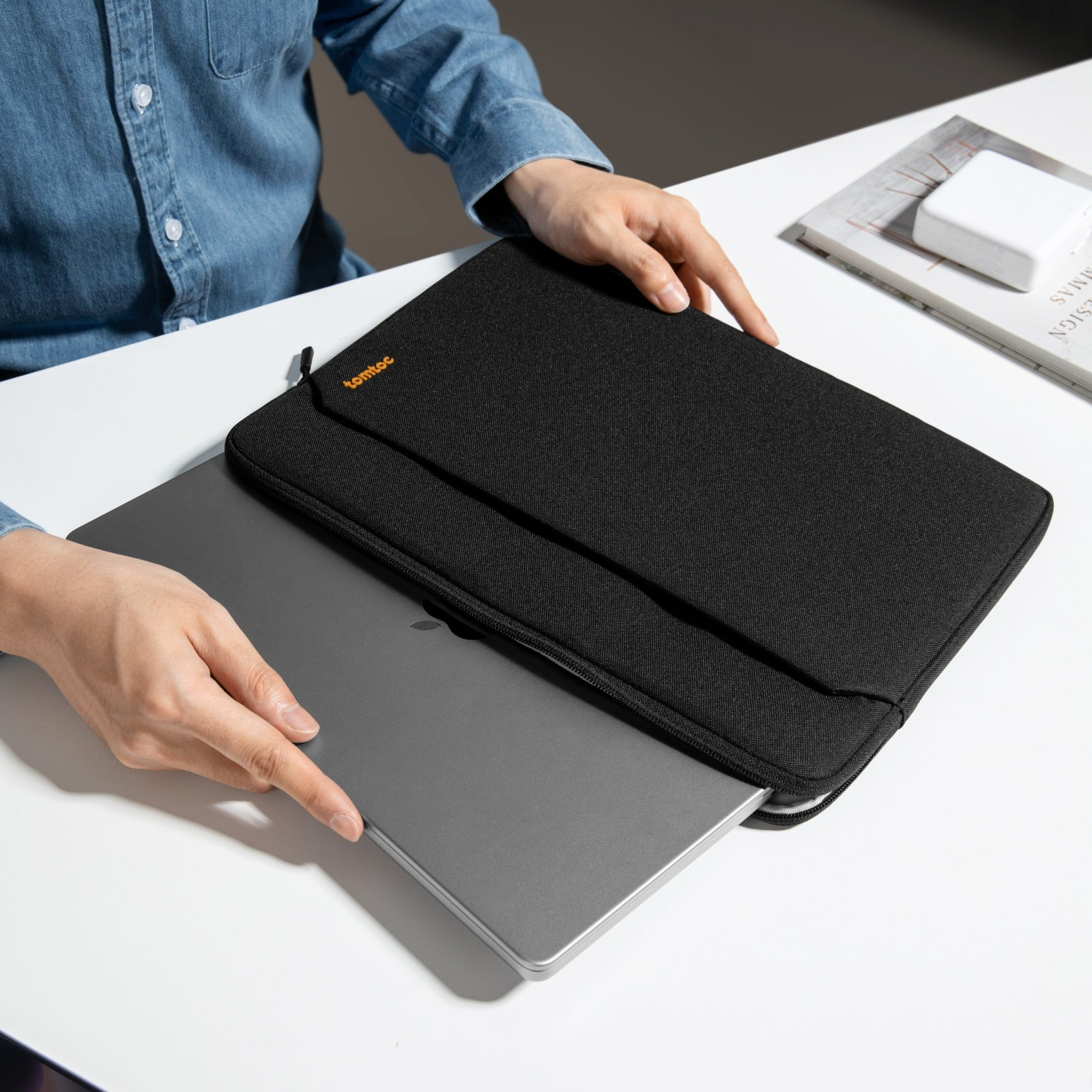 Osoba umieszczająca laptopa w czarnym etui na laptopa. Marka znajduje się na etui. Biała powierzchnia.