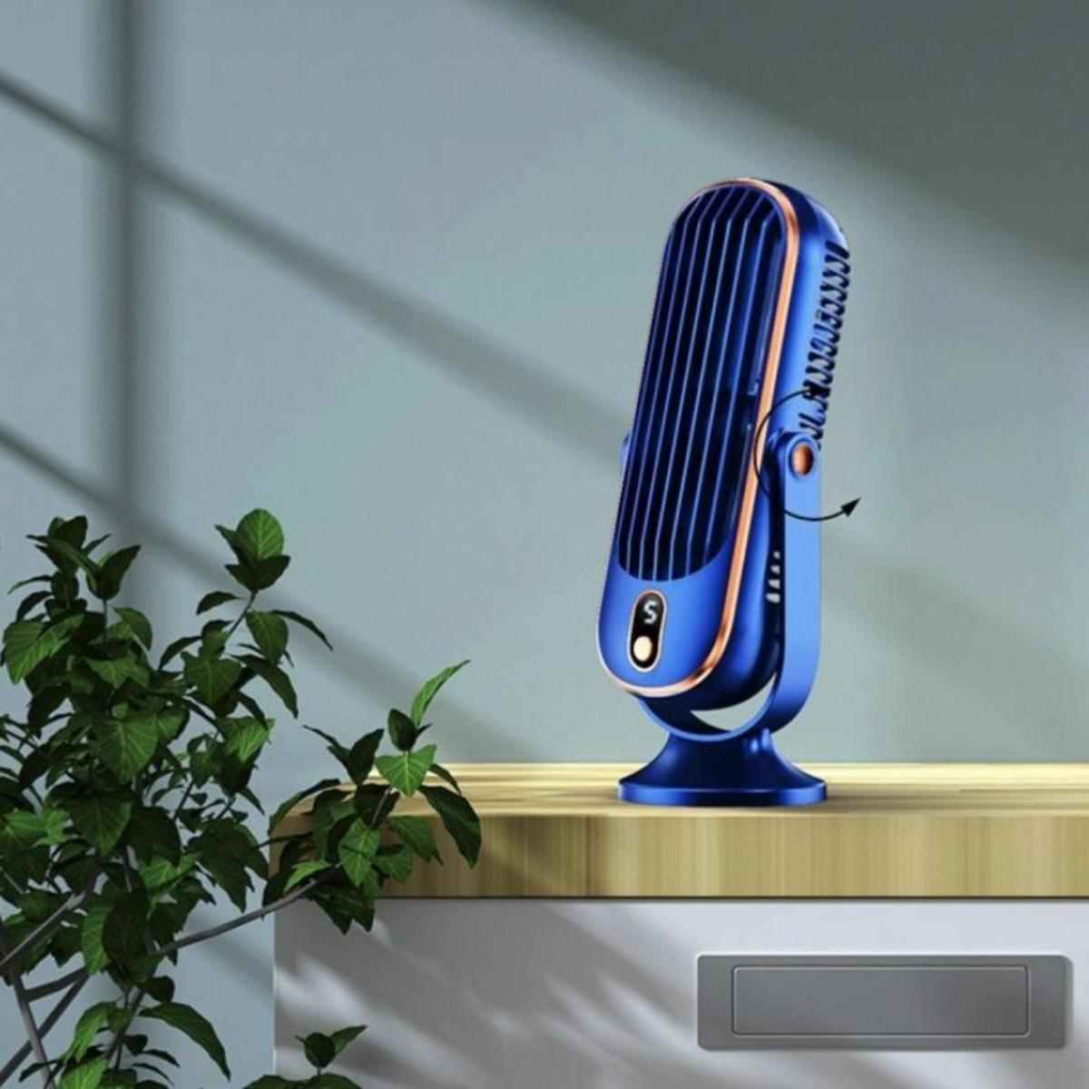 Ein blau-goldener Ventilator auf einer Holzoberfläche, links eine grüne Pflanze. Der Ventilator ist eingeschaltet.