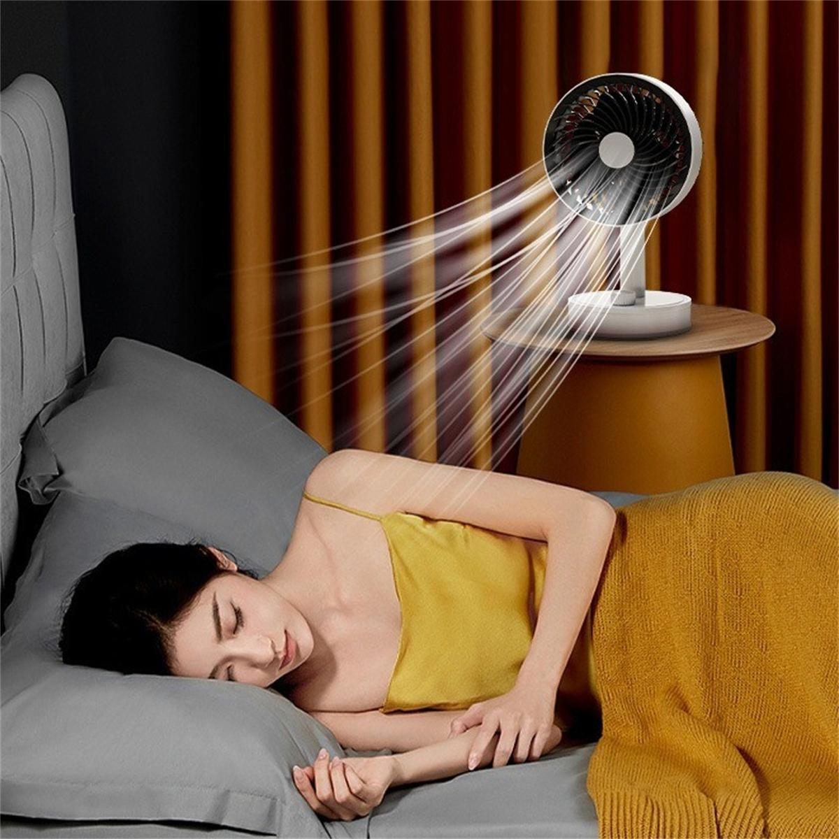 Eine Frau schläft im Bett, während ein Ventilator Luft auf sie bläst. Der Ventilator steht auf einem kleinen Tisch.
