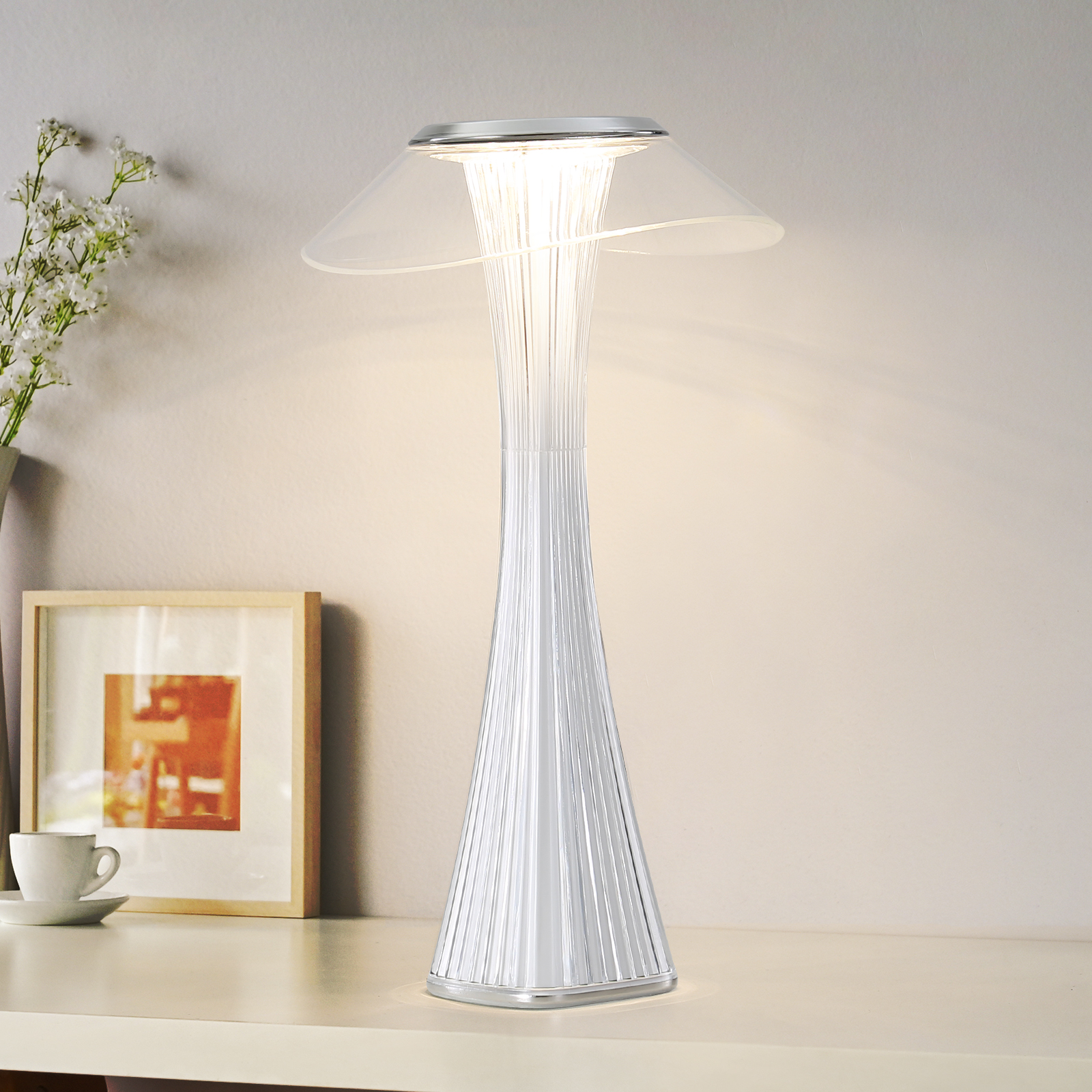 Lampa z przezroczystym, grzybkowatym blatem na białej powierzchni z ramką na zdjęcia i filiżanką.