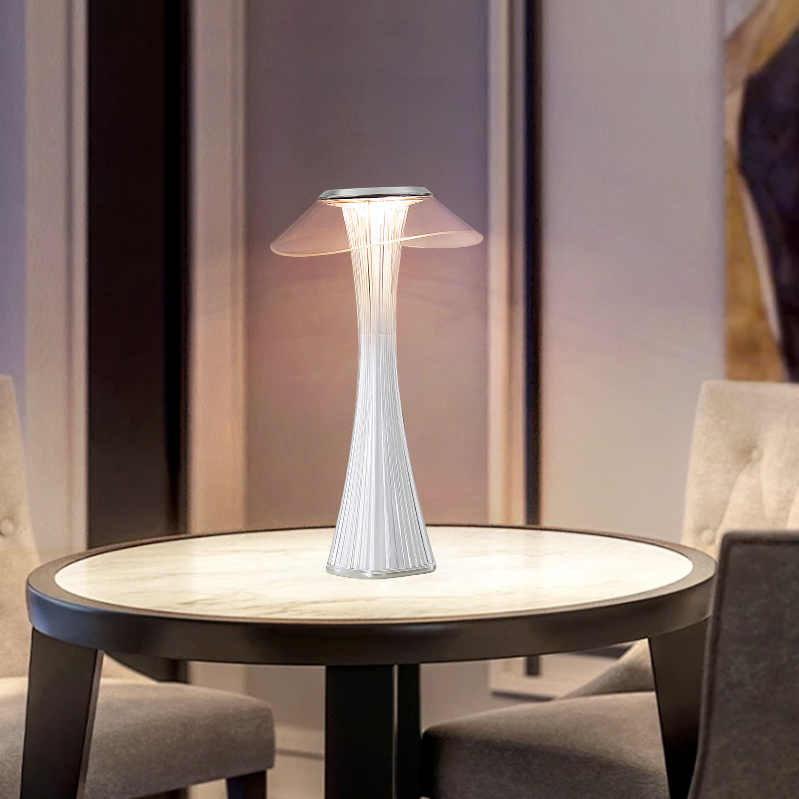 Biała lampa stołowa z przezroczystym kloszem. Jest zapalona i stoi na stole w pokoju.