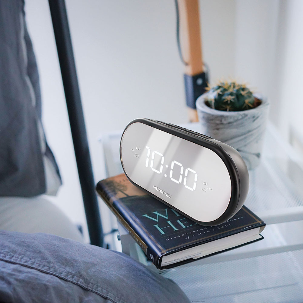Un reloj digital ovalado que muestra las 10:00. Está sobre un libro y una estantería con una planta al fondo.