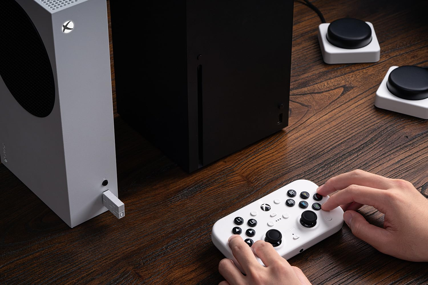 Manos sosteniendo un mando blanco conectado a una consola Xbox negra. Se ven dos botones adicionales.