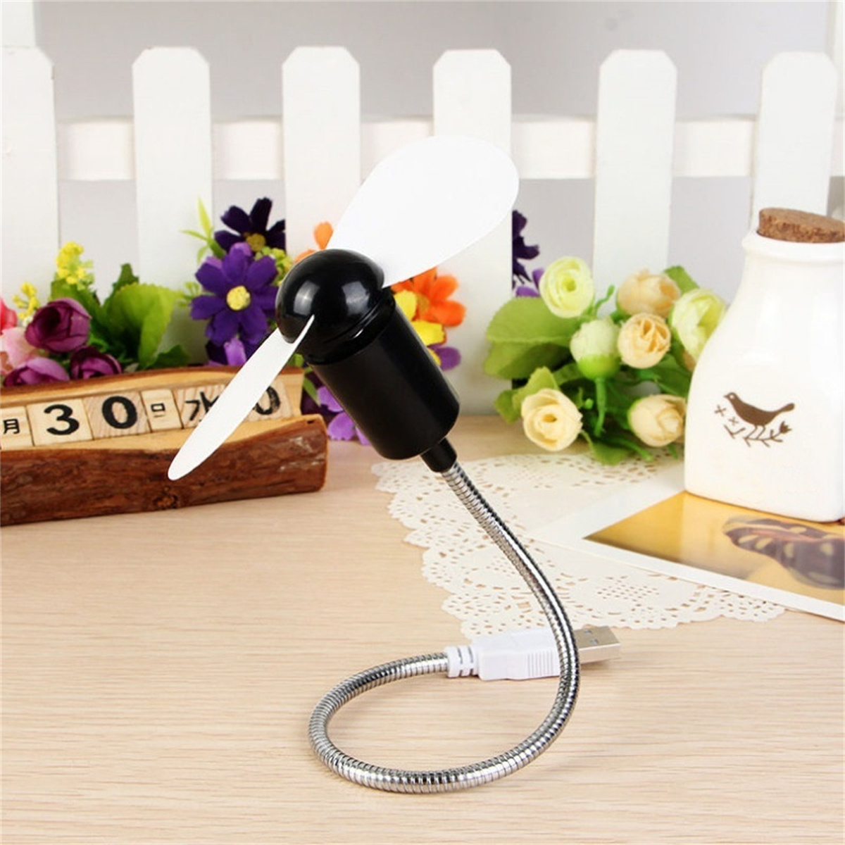 Ein schwarz-weißer USB-Ventilator steht auf einem Holztisch, im Hintergrund Pflanzen.