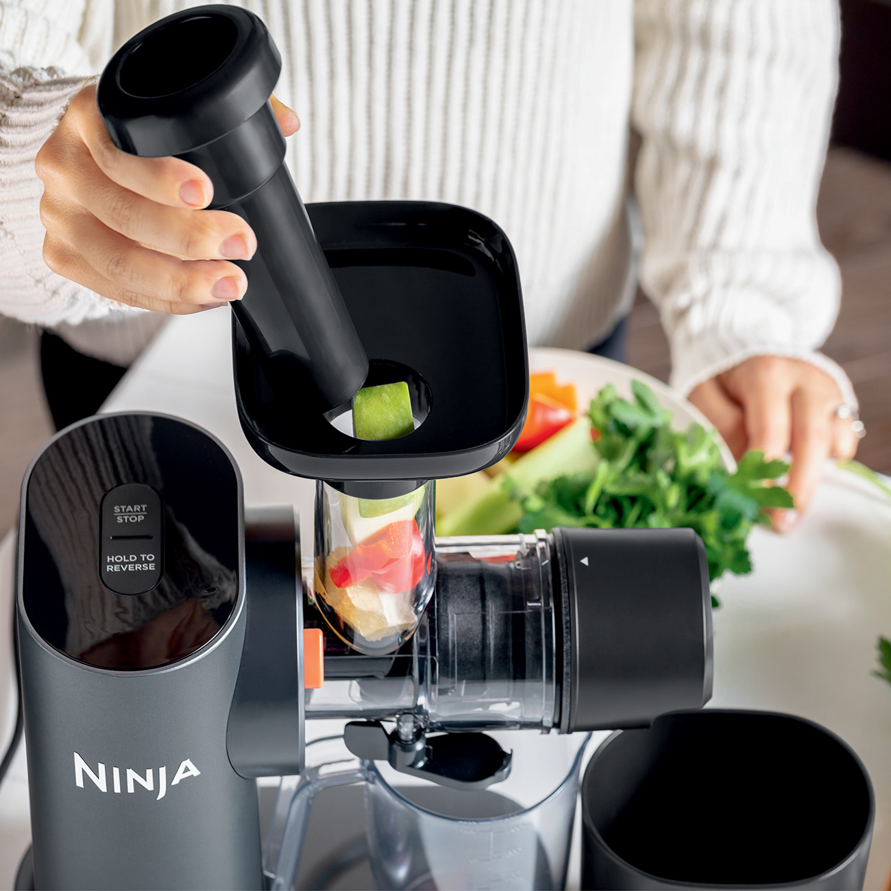 A person using a Ninja juicer. They are adding apple pieces. Other ingredients and a dish are on the counter.
