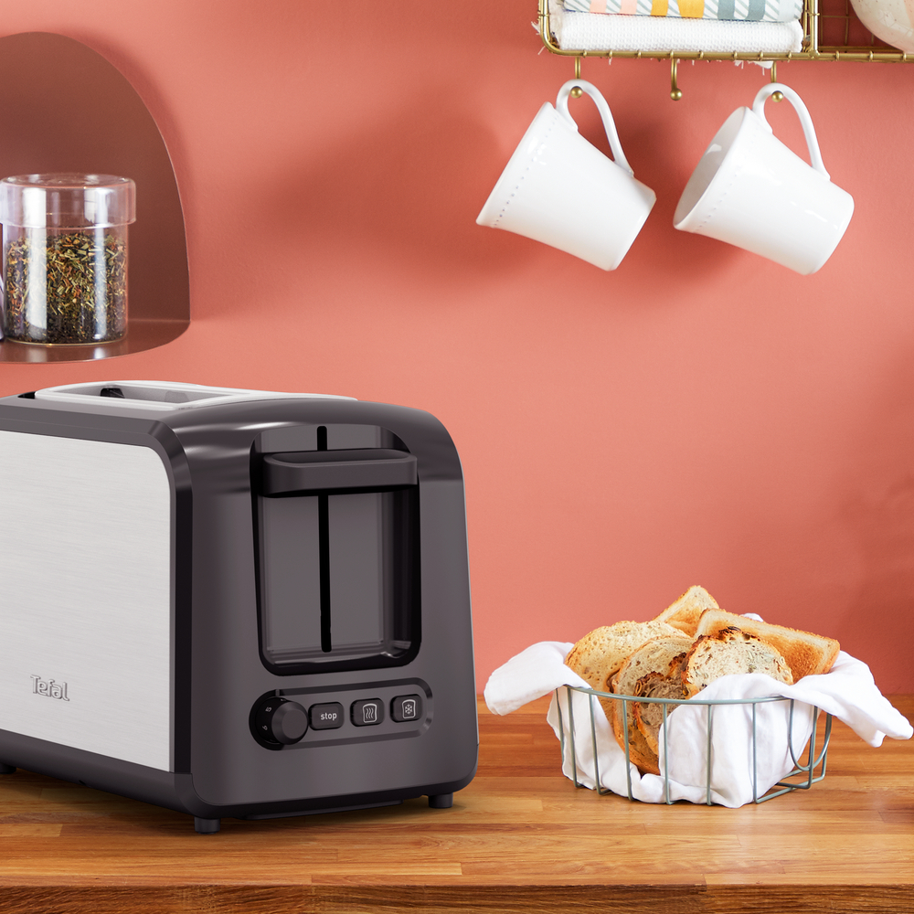 Toster obok kosza z chlebem i wiszących kubków, wszystko na drewnianym blacie.